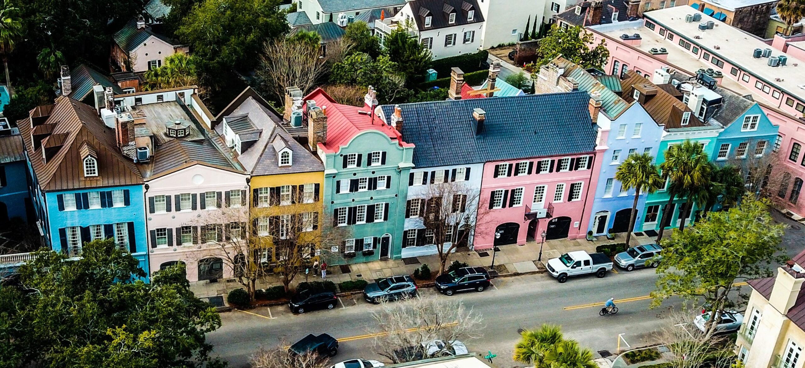 A row of colorful historic townhouses lines a street, each painted in pastel shades of blue, yellow, green, pink, and purple, with parked cars and a cyclist passing by on the road.