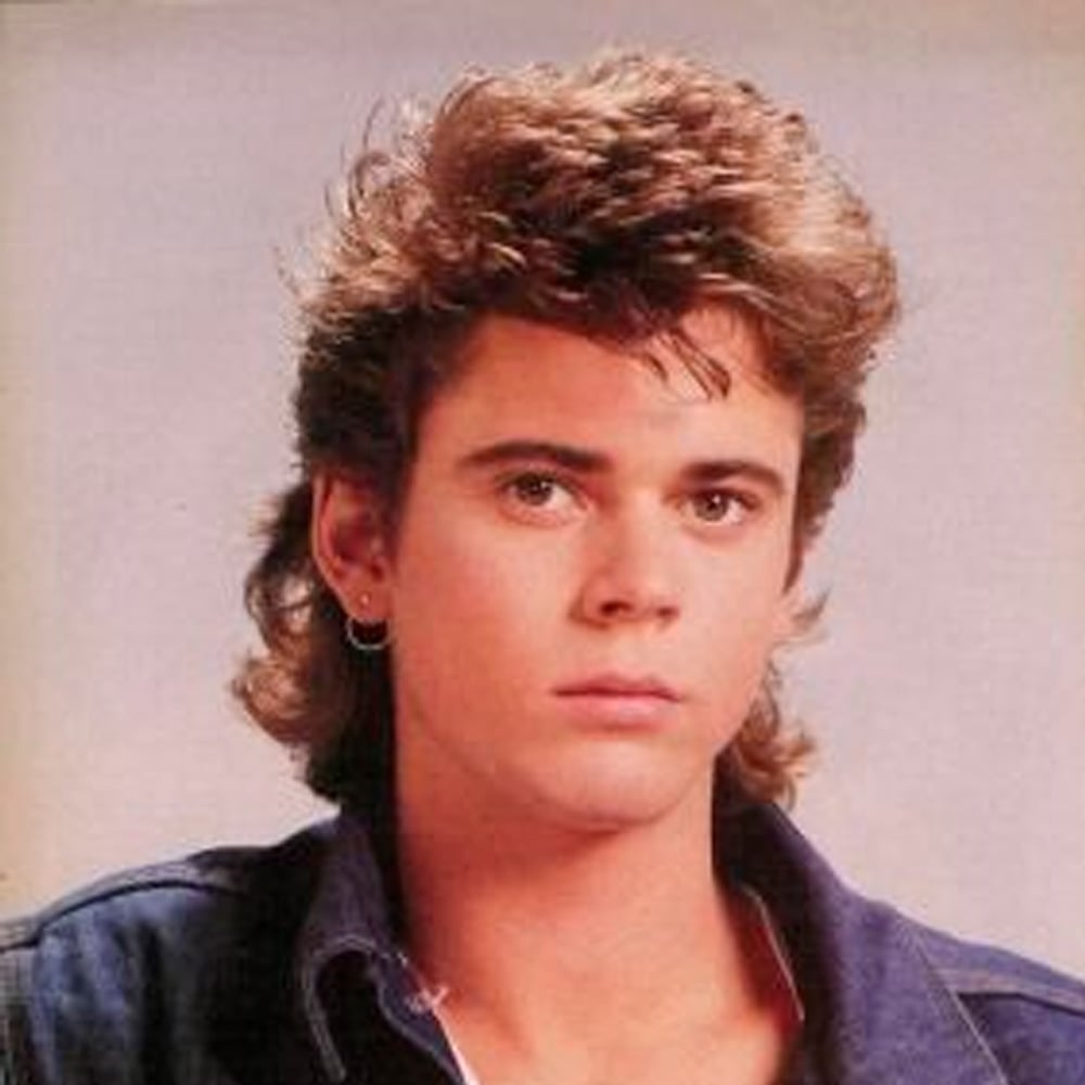 A young person with a mullet hairstyle, wearing a denim jacket and a small hoop earring, looks directly at the camera with a neutral expression against a plain background.