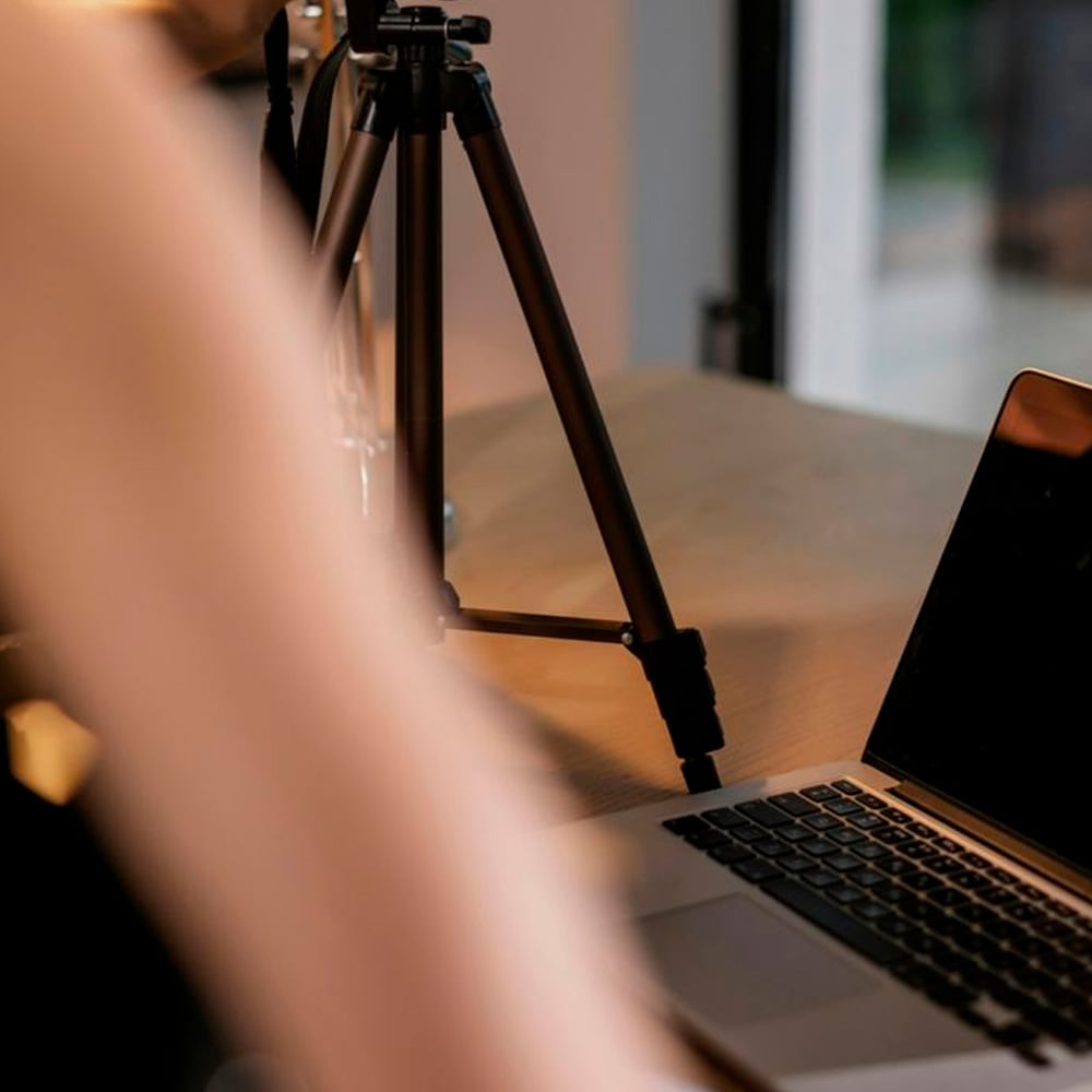 A close-up of a laptop on a wooden desk next to a camera tripod, with a blurred foreground and a window in the background.