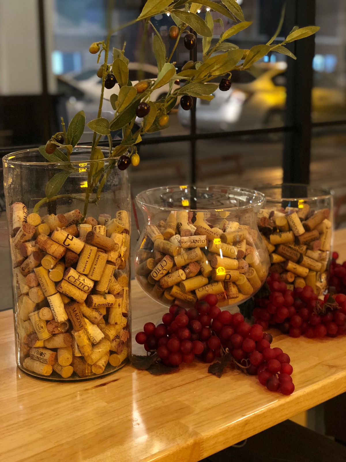 Three glass vases filled with wine corks are arranged on a wooden surface. Each vase has a branch with leaves and artificial grapes placed around them, creating a rustic wine-themed display.