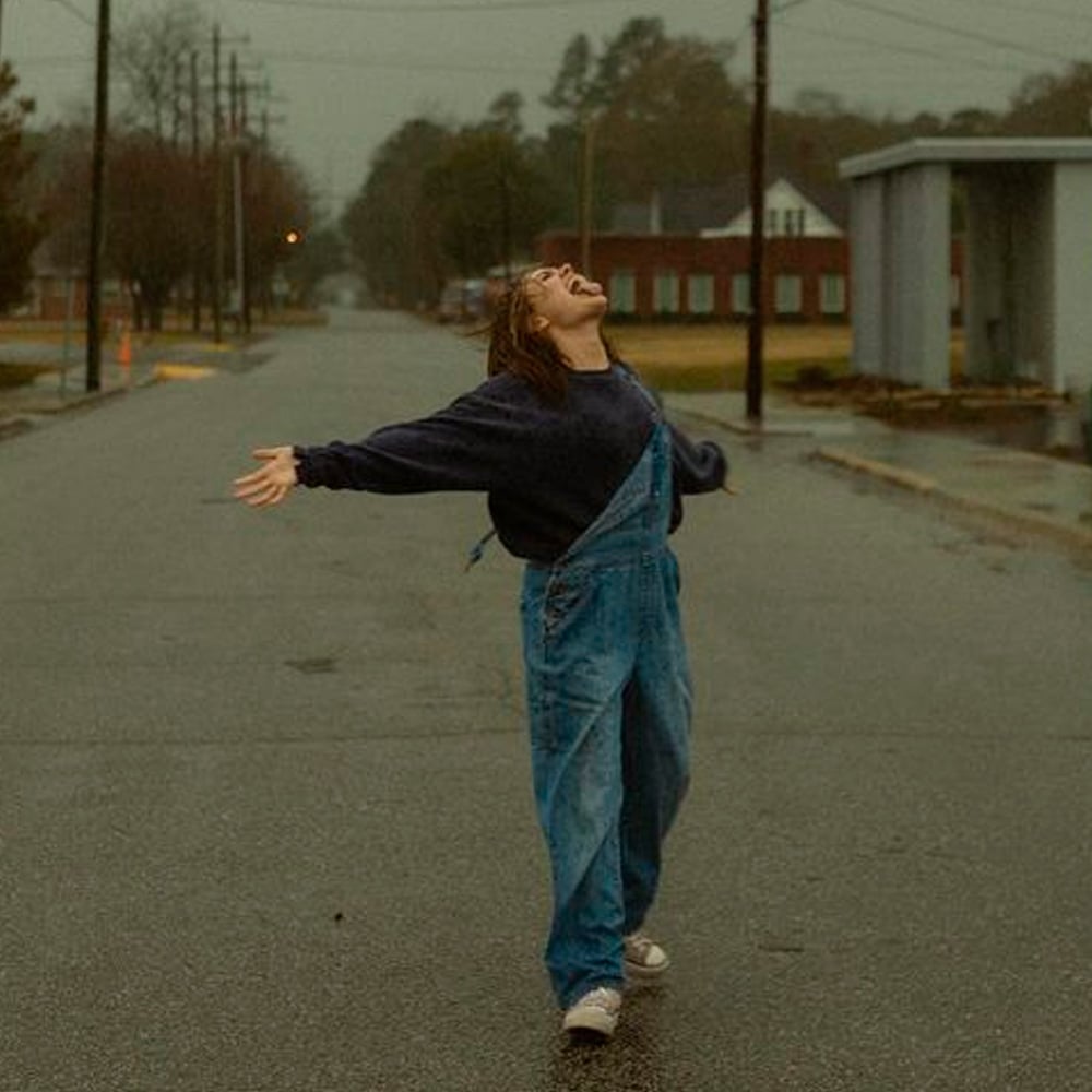 A person wearing blue overalls stands in the middle of a wet, empty street with arms outstretched and head tilted back, appearing joyful, on a cloudy, rainy day.