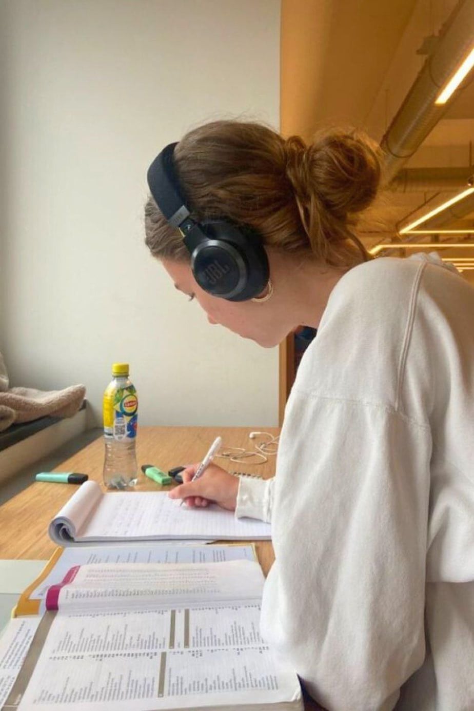 A person wearing headphones and a white sweater is studying at a desk with open notebooks, papers, a green highlighter, a pen, and a bottle of lemon iced tea. They are focused on writing or highlighting text.