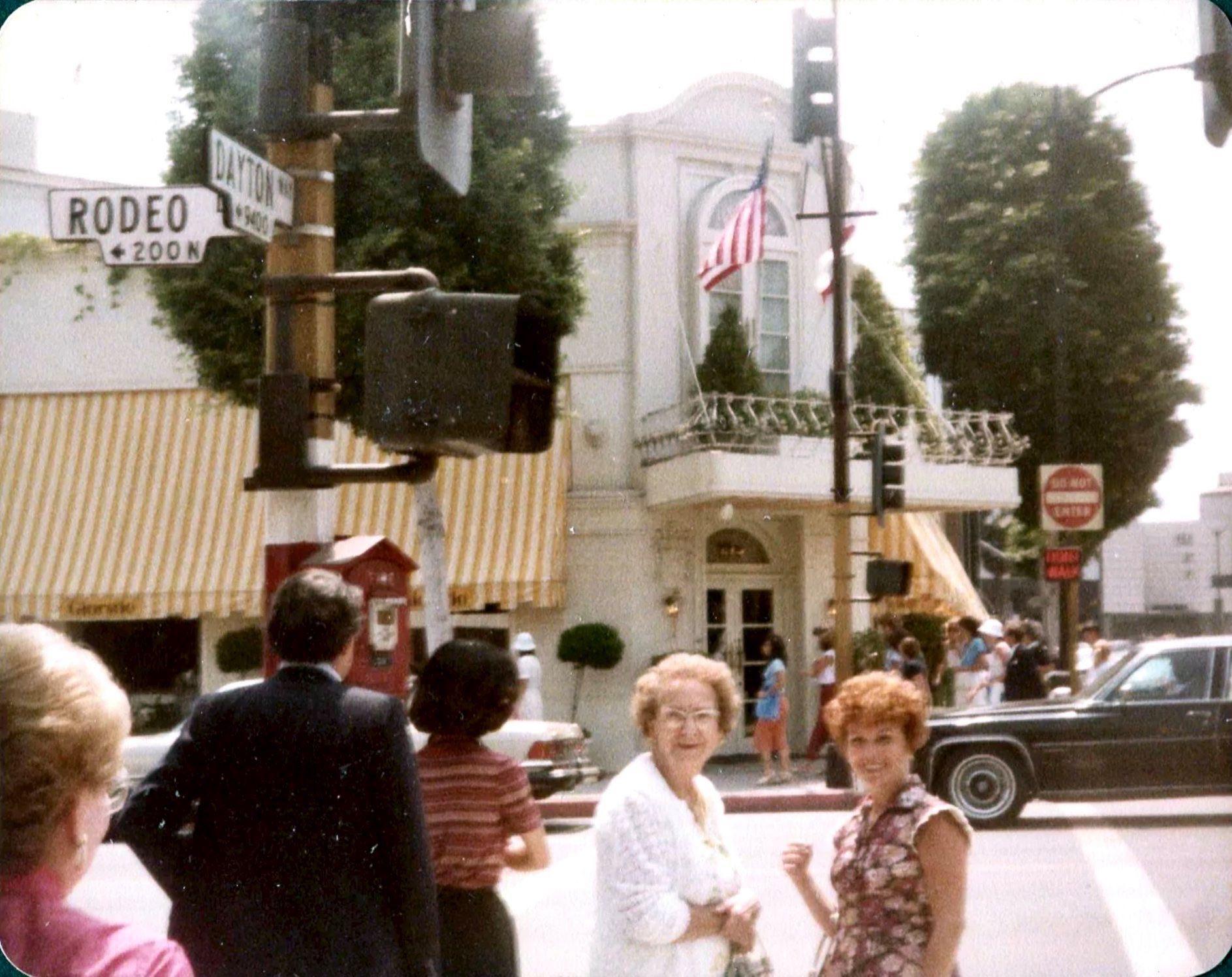 Two women stand on a busy street corner near a traffic light in front of a white building with yellow striped awnings. A street sign reads "Rodeo," and there are crowds, trees, and an American flag in the background.