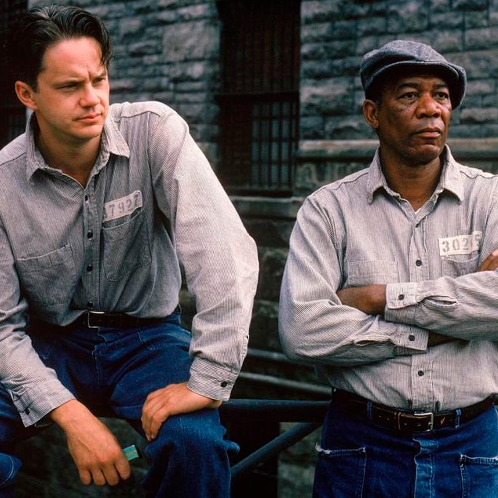 Two men wearing light gray prison uniforms sit and stand outside against a stone wall. One man has his arms crossed and wears a cap; the other is seated, both have serious expressions and prison number tags on their shirts.