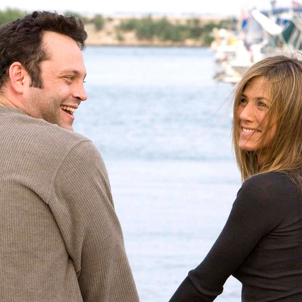 A man and woman stand by the water, smiling at each other. Boats and trees are visible in the background. Both are wearing long-sleeved shirts.