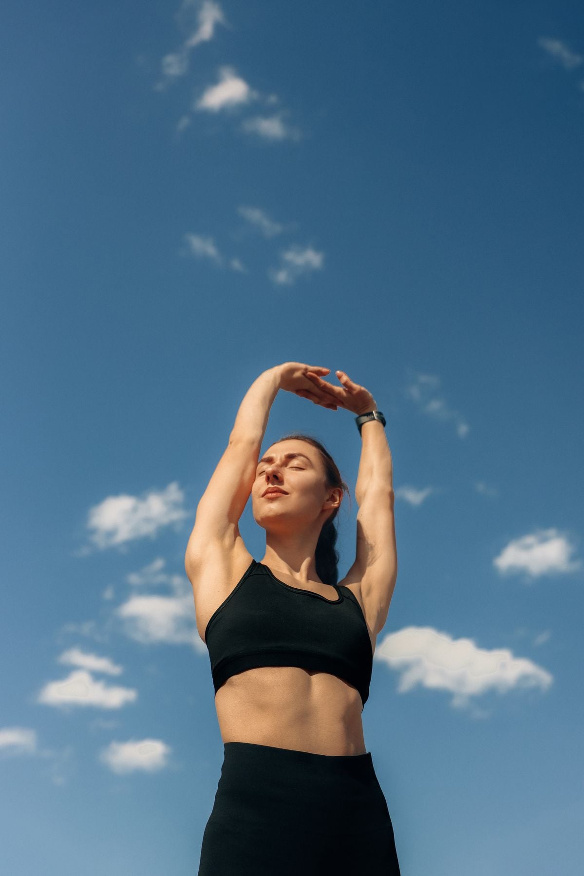 A woman in athletic wear stretches her arms overhead against a clear blue sky with scattered white clouds, looking relaxed and focused.