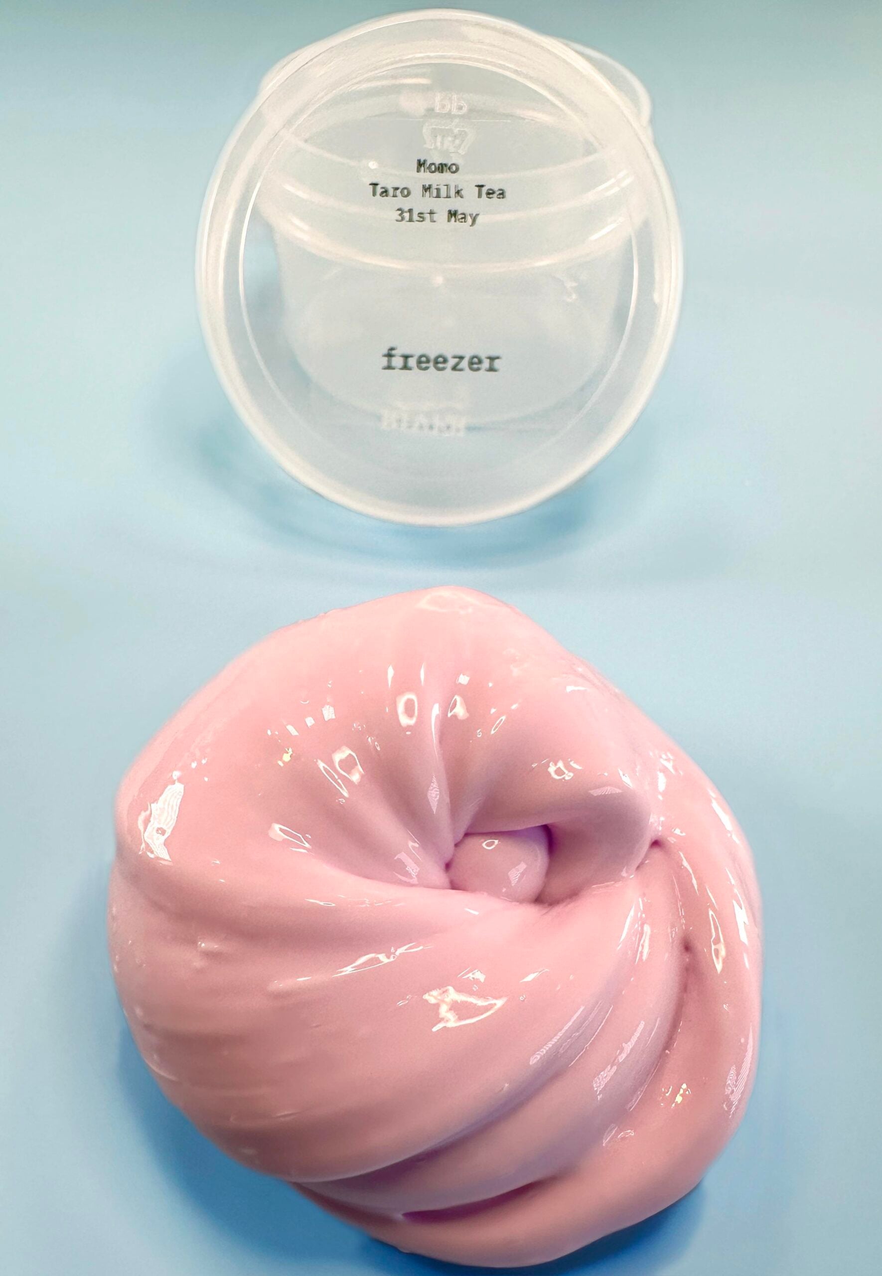 A glossy, light pink slime is swirled on a light blue surface, with a transparent container labeled “Mono Taro Milk Tea 31st May freezer” placed above it.