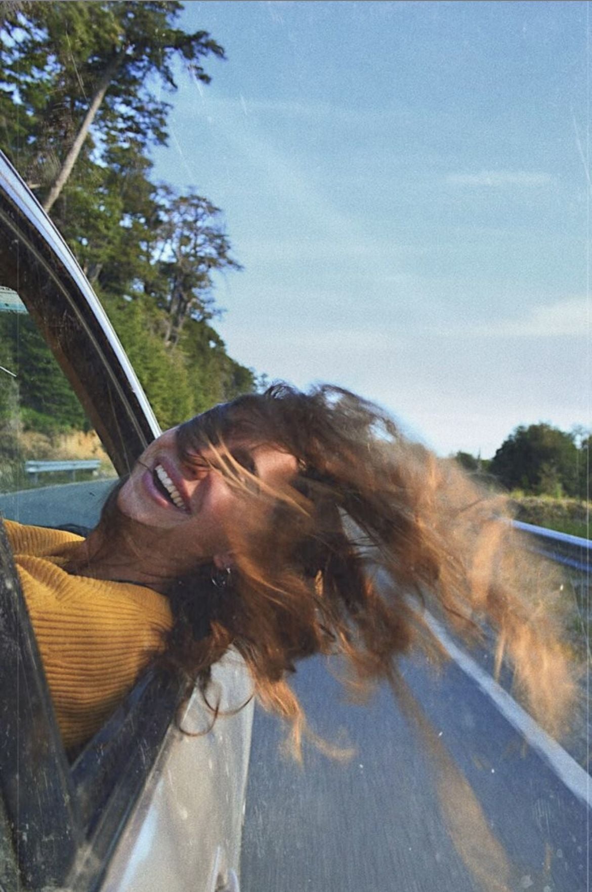 A person with long, wavy hair leans out of a car window, smiling joyfully as their hair blows in the wind on a sunny day. The car drives along a road bordered by trees and blue sky.