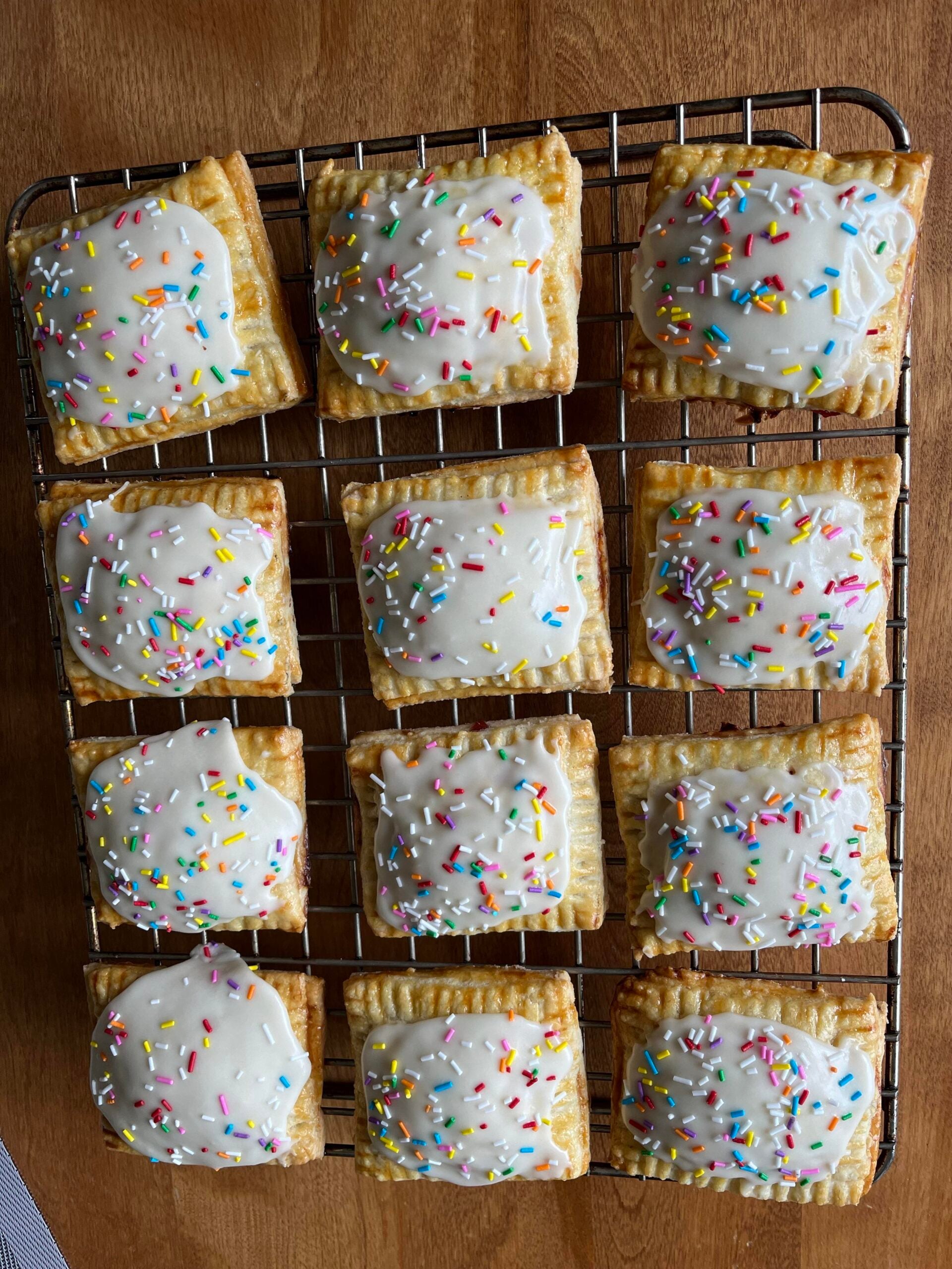 Twelve homemade rectangular pastries with white icing and colorful sprinkles are cooling on a wire rack over a wooden surface.