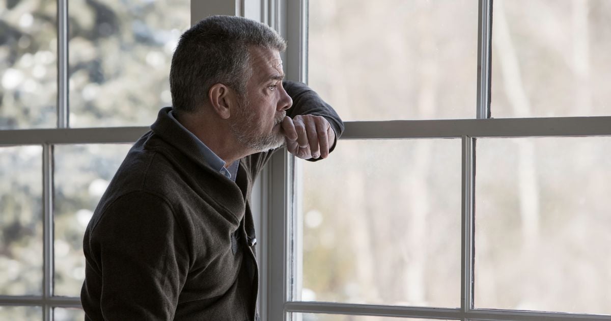 A middle-aged man with gray hair and a beard looks thoughtfully out a large window, resting his chin on his hand. He is indoors, wearing a dark sweater, with a blurred outdoor scene in the background.