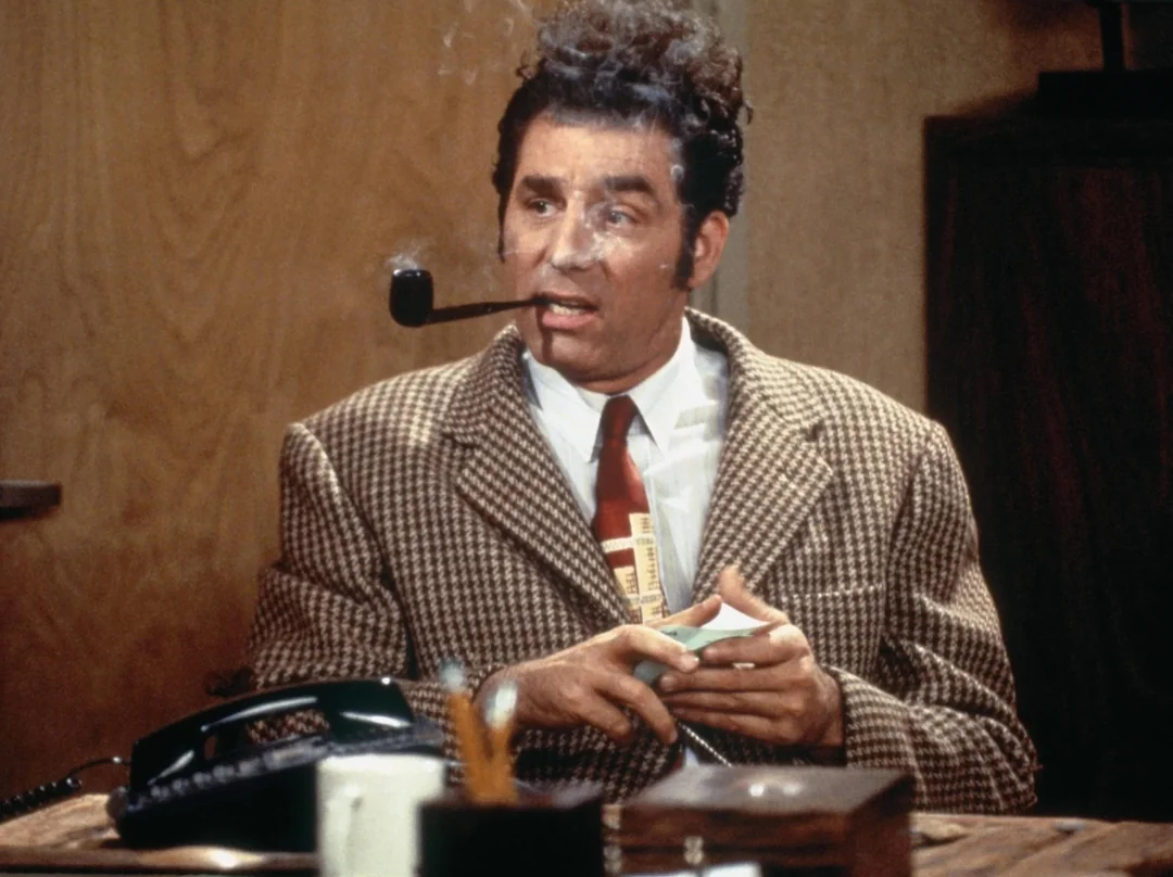 A man in a checkered jacket and tie sits at a desk holding papers, with a pipe in his mouth. His hair is messy and he appears surprised. There is an old-fashioned telephone and a mug on the desk.