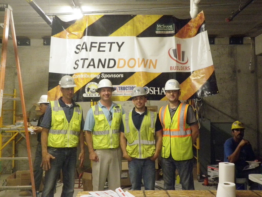 Four construction workers in safety vests and hard hats stand and smile in front of a "Safety Stand Down" banner at a construction site. More workers and site equipment are visible in the background.