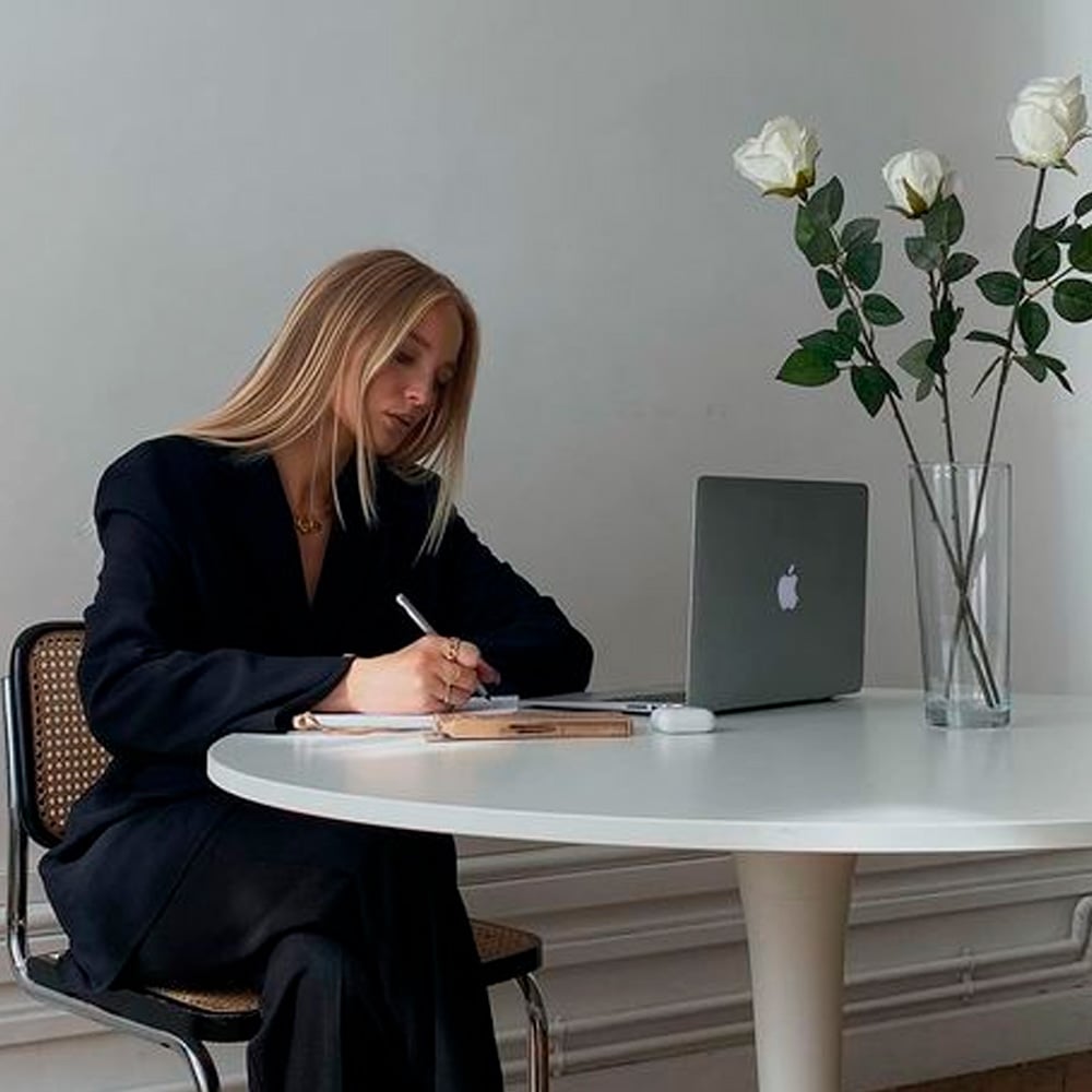 A person with long blonde hair sits at a round white table, writing in a notebook beside an open laptop. A vase with white roses is placed on the table near the computer.