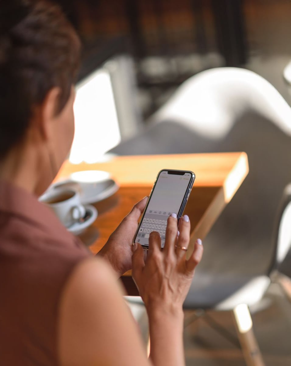 A person sits at a table in a café, holding and typing on a smartphone. A cup of coffee and a glass of water are on the table beside them. The focus is on their hands and phone.