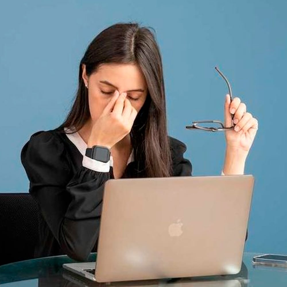 A woman sitting at a desk in front of a laptop holds her glasses in one hand while pinching the bridge of her nose, appearing tired or stressed.