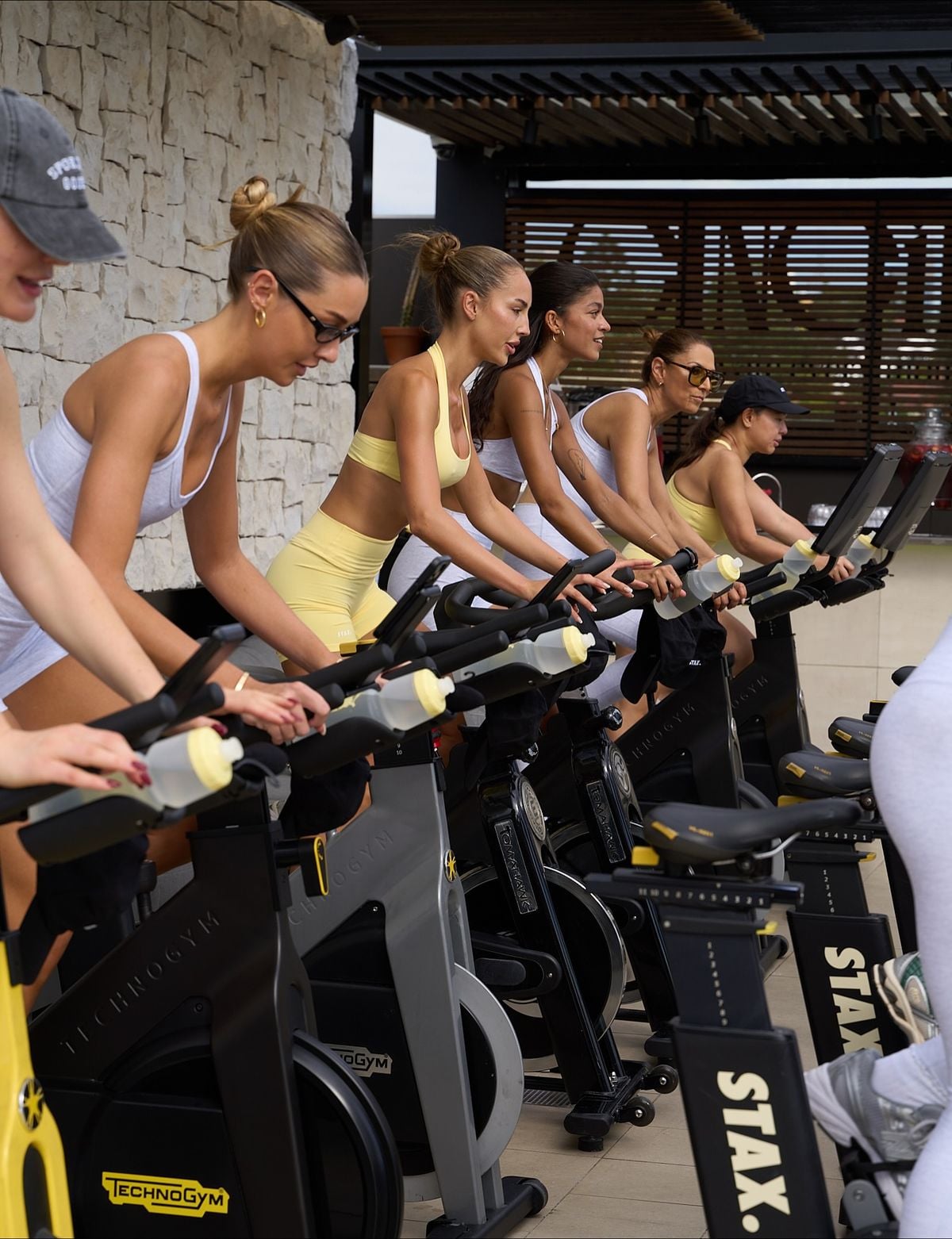 A group of women in activewear ride stationary bikes outdoors, exercising side by side in a row. The setting appears to be a sunny, modern patio with stone and wood elements in the background.