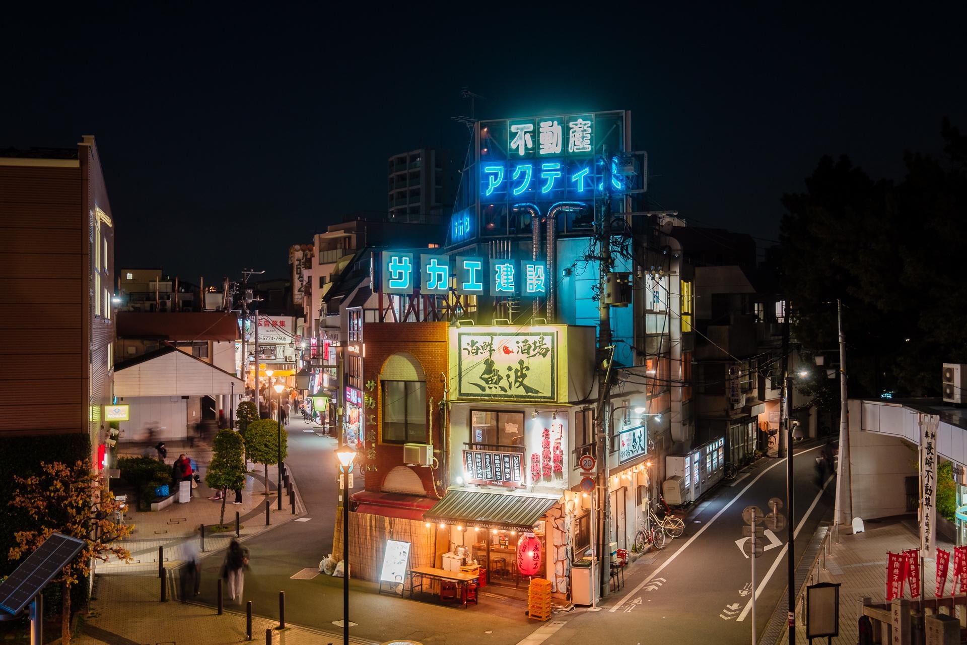 A lively Japanese street at night with illuminated neon signs, restaurants, and shops. People walk along the sidewalks, and the scene is brightly lit with colorful lights and signage.