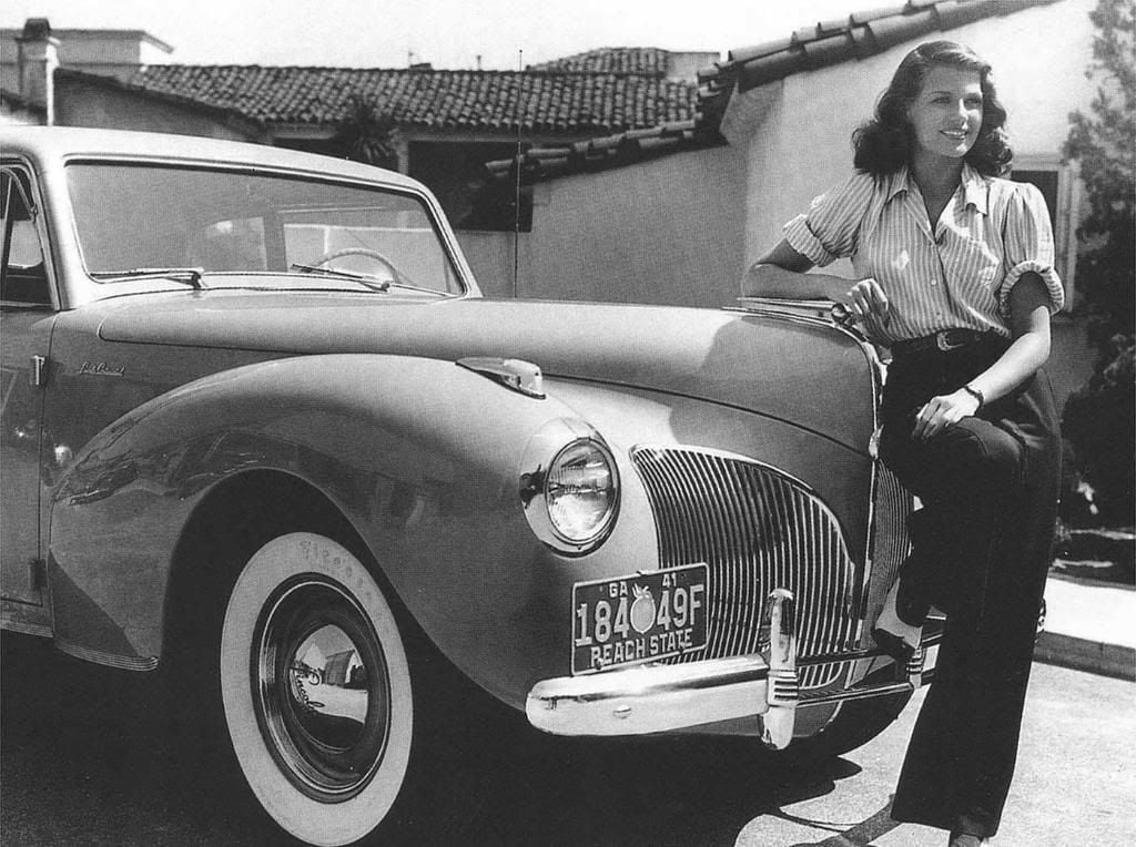 A smiling woman in a striped blouse and dark pants leans against the front of a vintage car parked in a driveway, with a house and trees visible in the background. The car has a visible 1940s license plate.