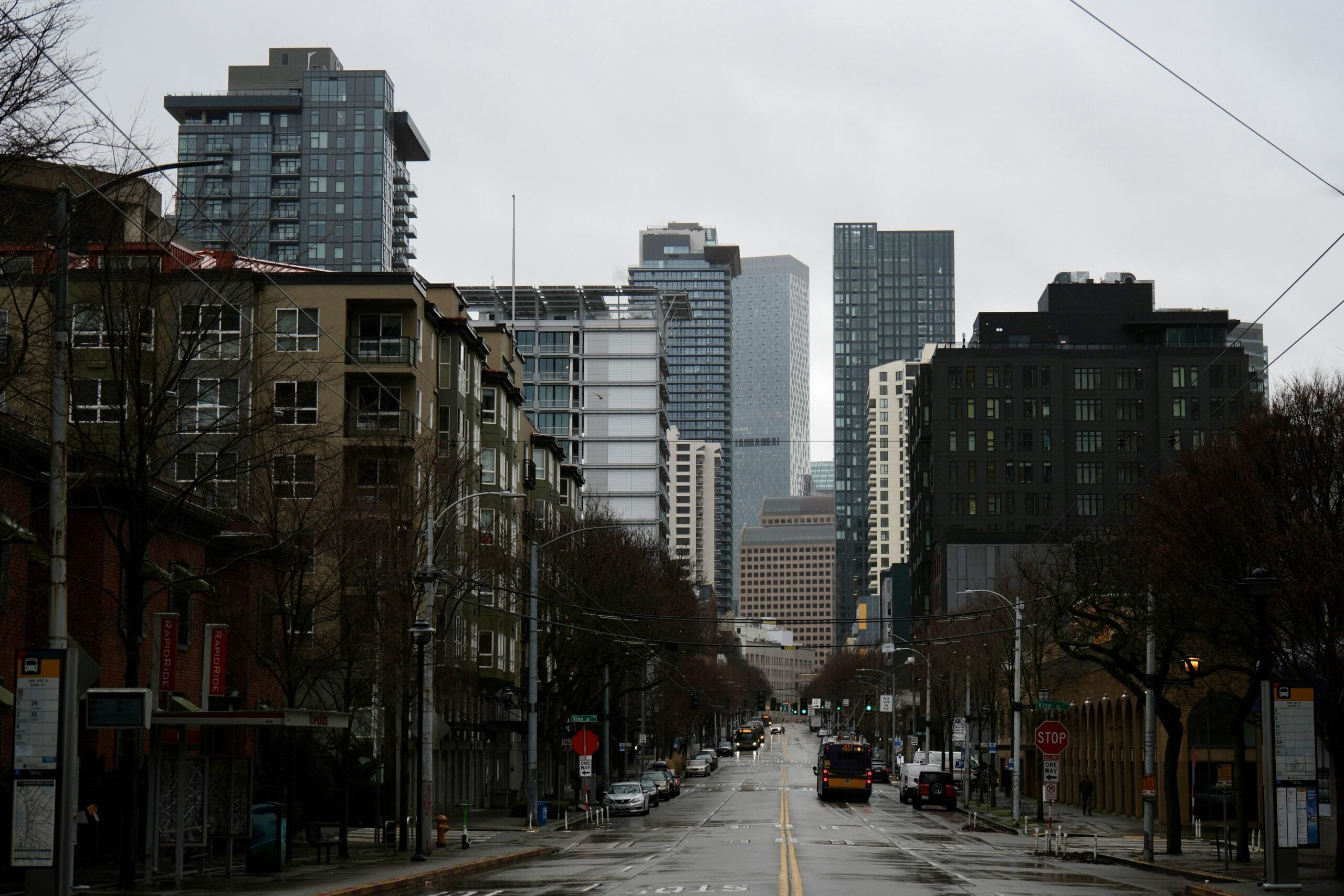 A city street lined with buildings on both sides leads toward tall skyscrapers in the background on a cloudy, overcast day. There are a few cars, buses, and bare trees along the sidewalks.