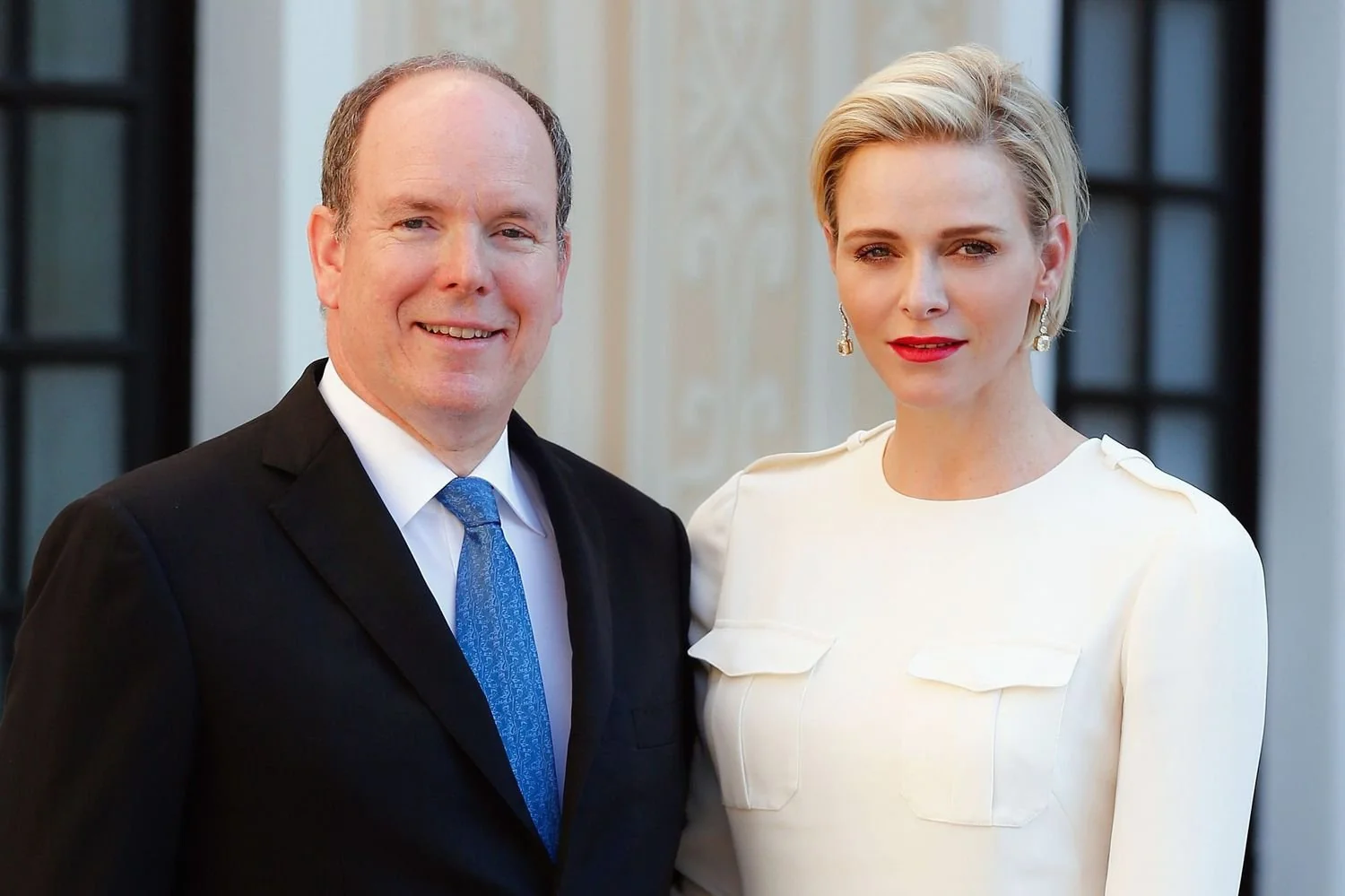 A man in a dark suit and blue tie stands next to a woman in a white dress with earrings, both smiling slightly and posing for a formal photo in front of an elegant building.