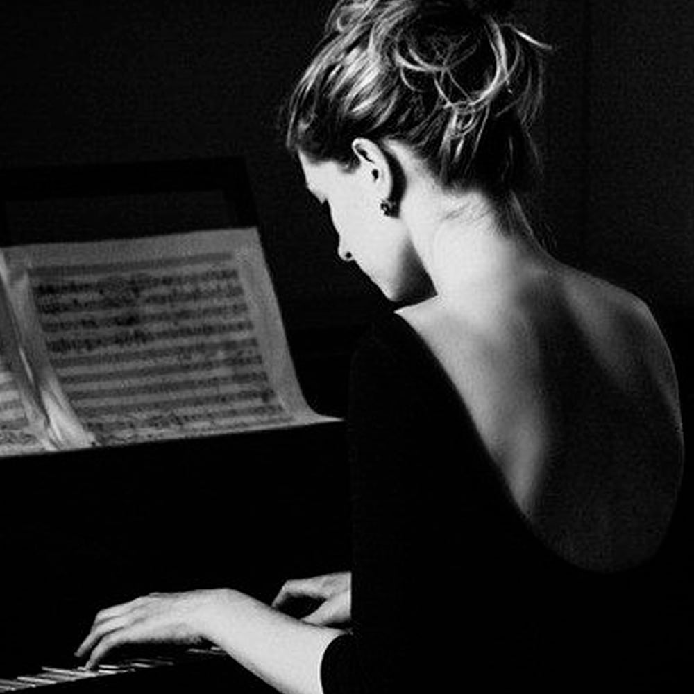 A woman with her hair in a loose bun and wearing a backless dress plays the piano, with sheet music propped up in front of her. The image is in black and white.