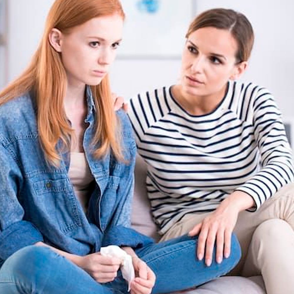 A concerned woman comforts a sad young woman sitting beside her on a couch. The young woman holds a tissue and looks away, while the older woman rests a reassuring hand on her shoulder.