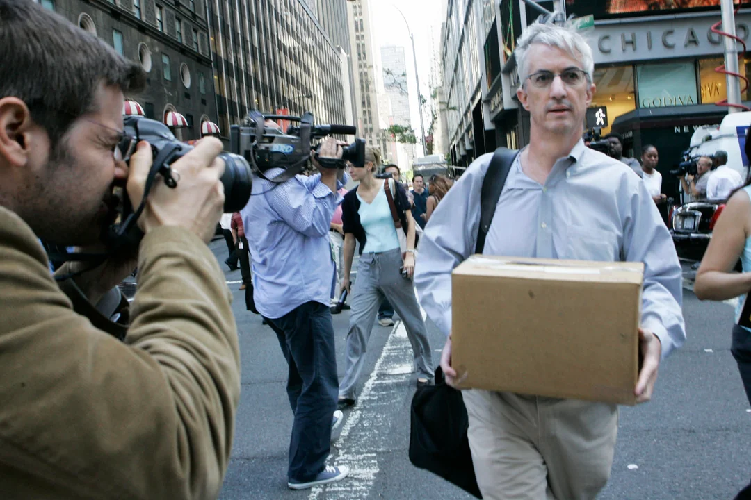 A man with gray hair and glasses walks down a busy city street carrying a cardboard box, while people around him take photos and record video. Tall buildings and storefronts line the street in the background.