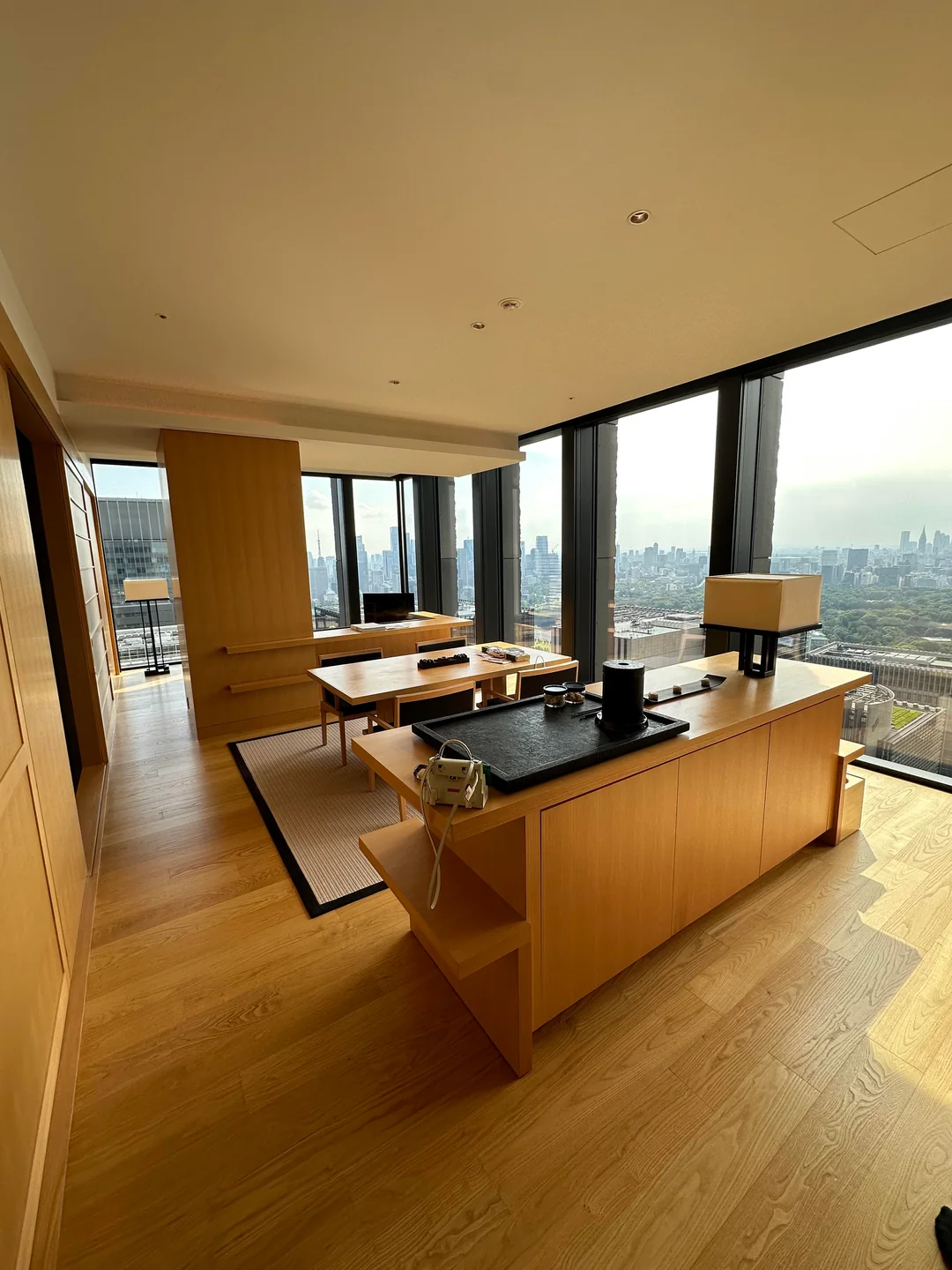 A modern, sunlit hotel room with wooden floors, minimalist furniture, large windows, and a city skyline view. A desk with a lamp and phone sits in the foreground, and a table with chairs is near the windows.