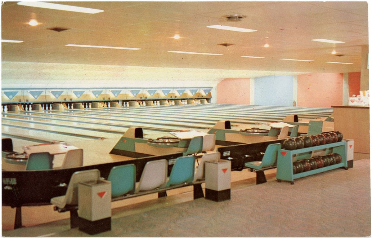 A vintage bowling alley with multiple empty lanes, retro seating, and ball returns. Bowling balls are lined up on racks, and the area is brightly lit with overhead lights. No people are present.