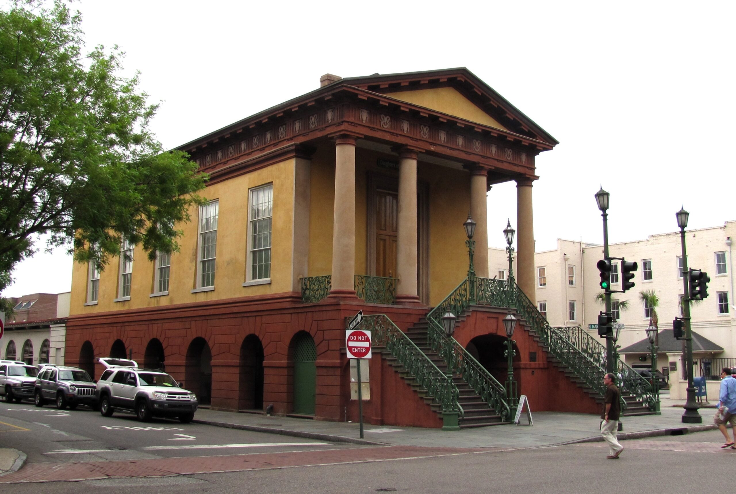 A historic two-story building with large columns, tall windows, and a wide staircase with green railings, situated on a street corner with parked cars and a pedestrian nearby.