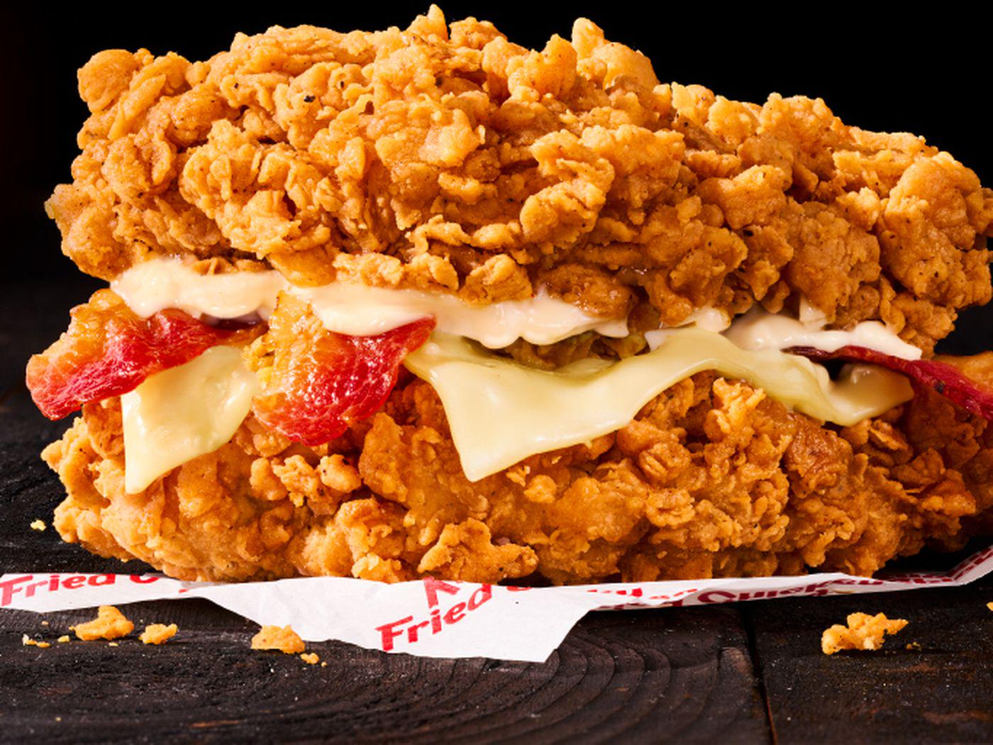 Two crispy fried chicken fillets serve as the "bun," sandwiching slices of melted cheese, bacon, and sauce, all set on a piece of branded paper against a dark background.