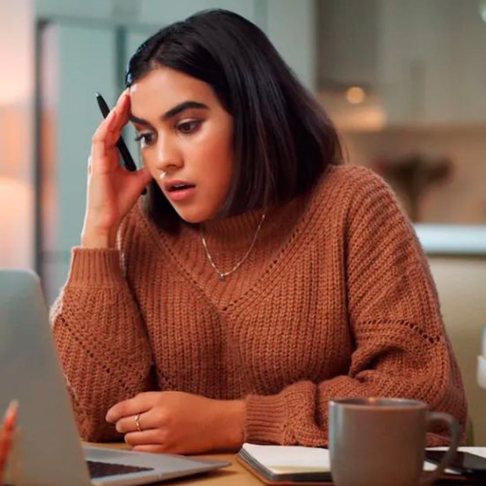 A young woman with dark hair wearing a brown sweater looks surprised or stressed while holding a pen and staring at a laptop screen, with a notebook and mug on the table in front of her.