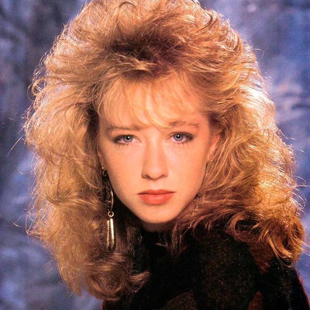 A woman with voluminous, curly blonde hair styled in an 80s fashion, wearing a dark top and long, dangling earrings, looks directly at the camera with a neutral expression against a soft, bluish background.