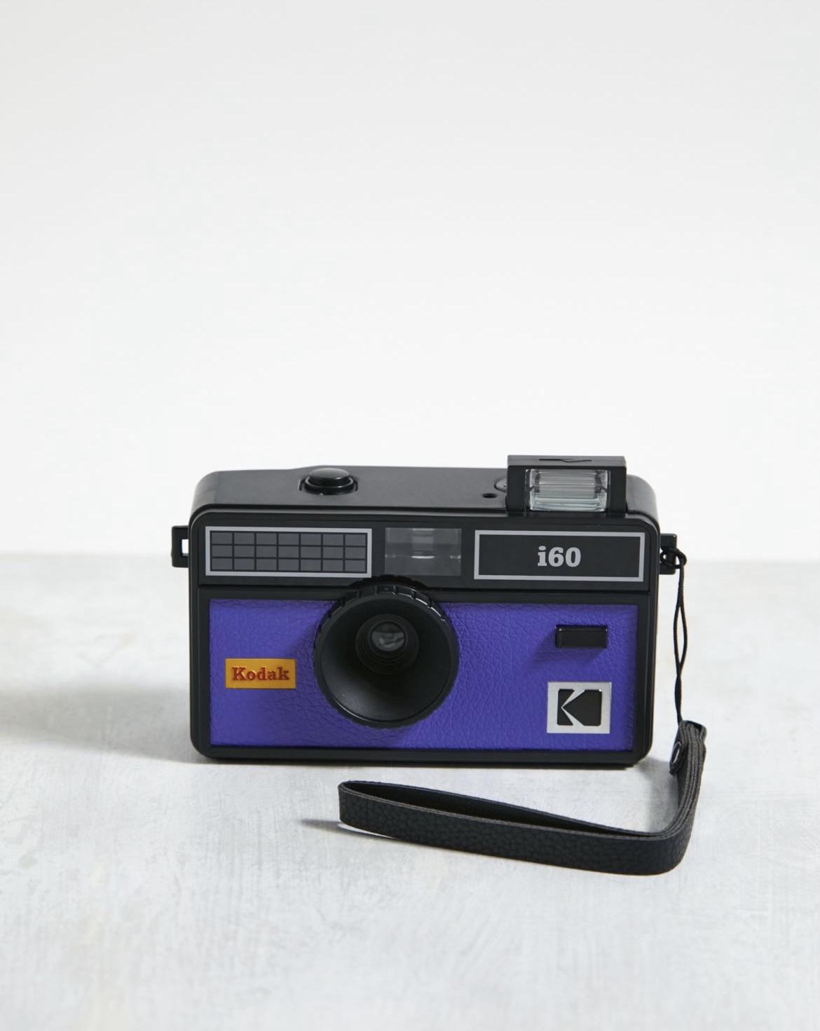 A Kodak i60 film camera with a purple front, black lens, and black wrist strap sits on a light-colored surface against a plain white background.