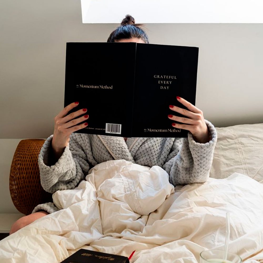 A person in a gray robe sits in bed under a white blanket, holding and reading a book titled "Grateful Every Day." Their face is mostly hidden by the book.