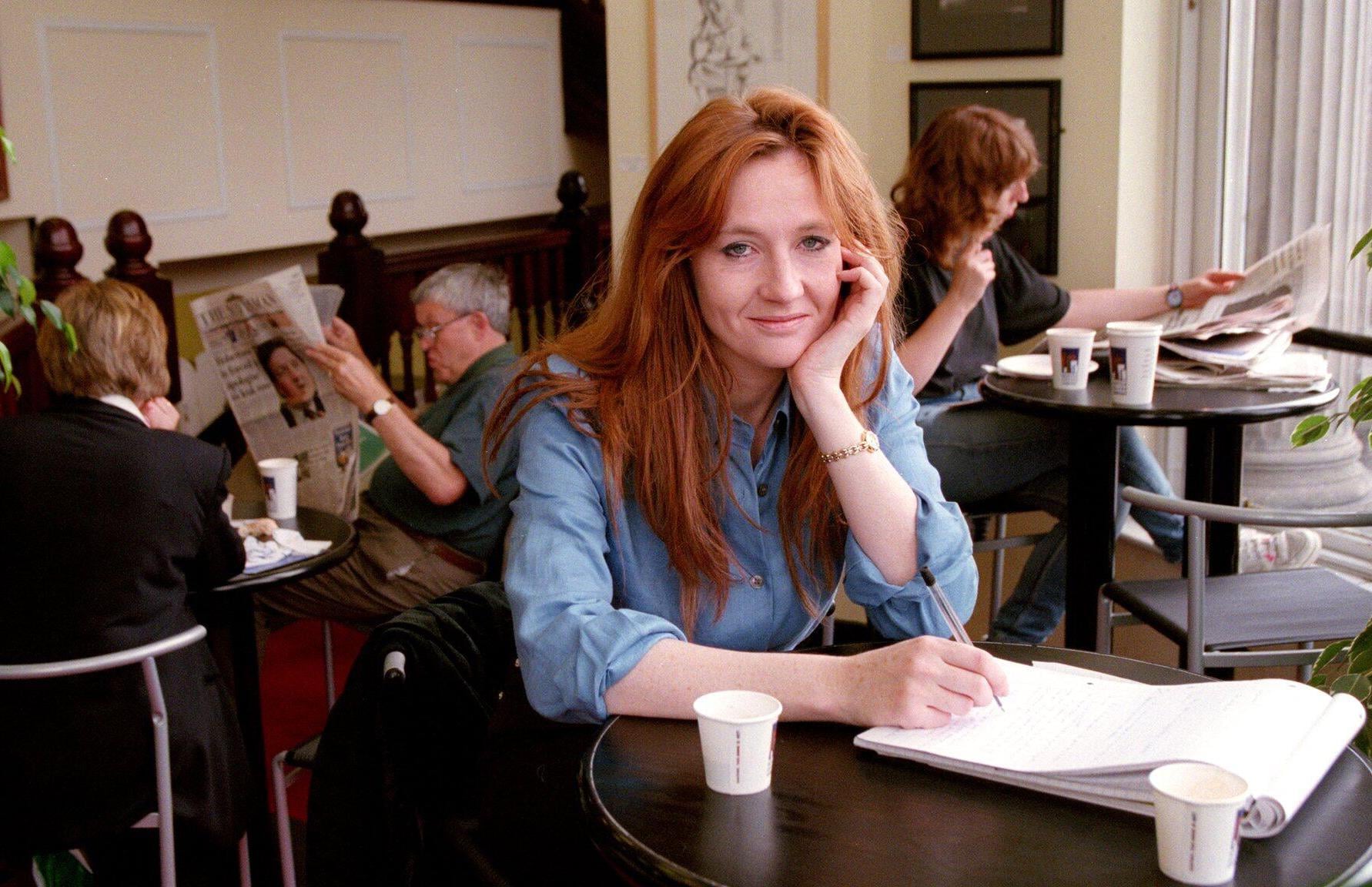 A woman with long red hair, wearing a blue shirt, sits at a table with papers and a coffee cup, smiling at the camera in a café. Other people are reading and drinking coffee in the background.