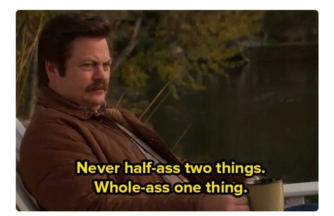 A man with a mustache sits outdoors by a lake, holding a mug. Text on the image reads: "Never half-ass two things. Whole-ass one thing.