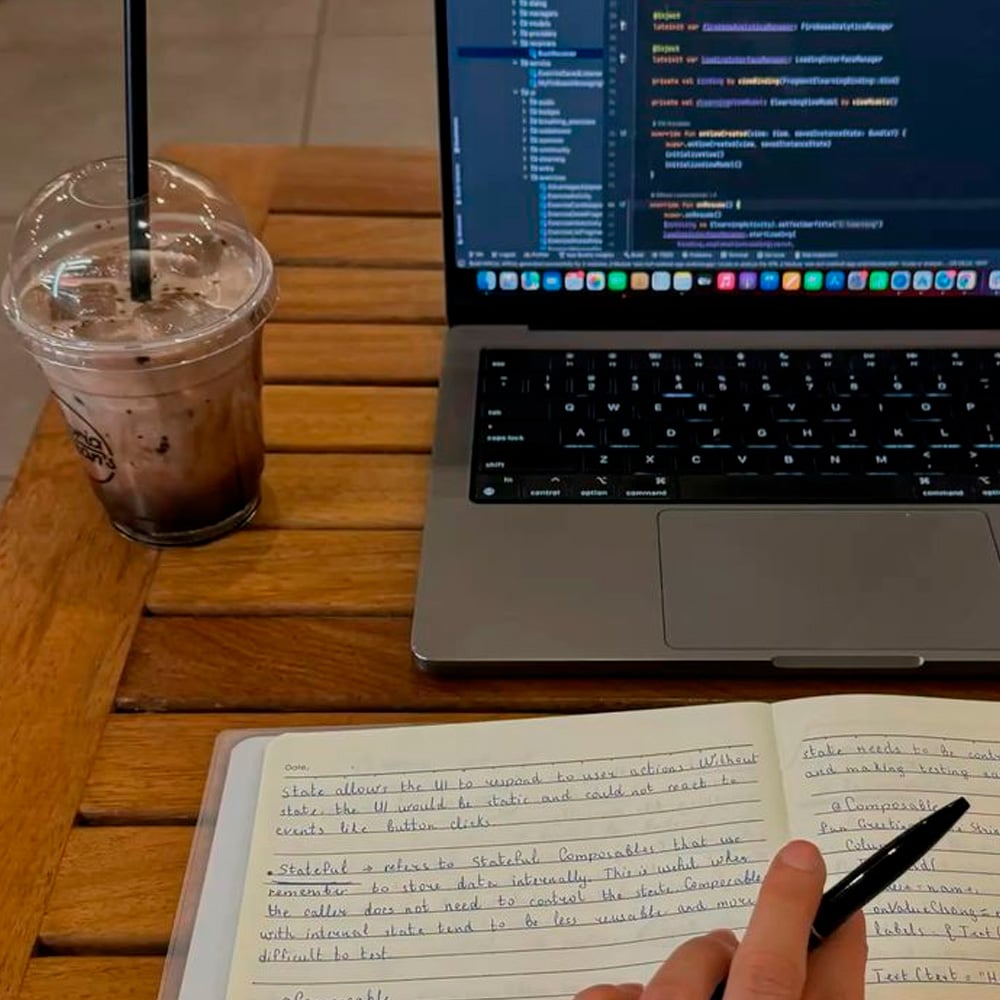 A person writes in a notebook with handwritten notes, while a laptop with code on the screen sits on a wooden table. There is also an iced drink with a straw next to the laptop.