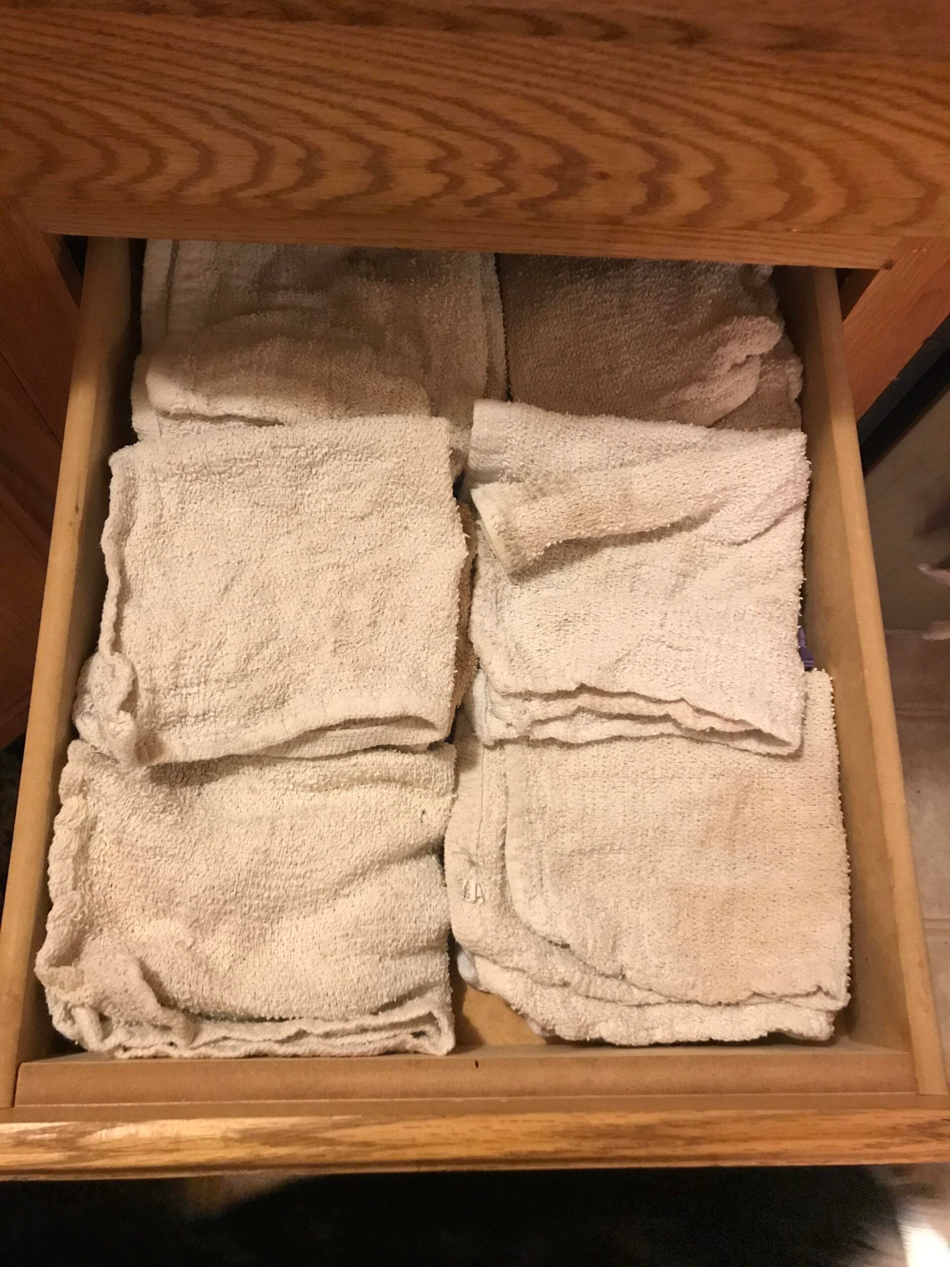 A wooden drawer partially open, filled with neatly folded off-white washcloths or rags arranged in two rows.