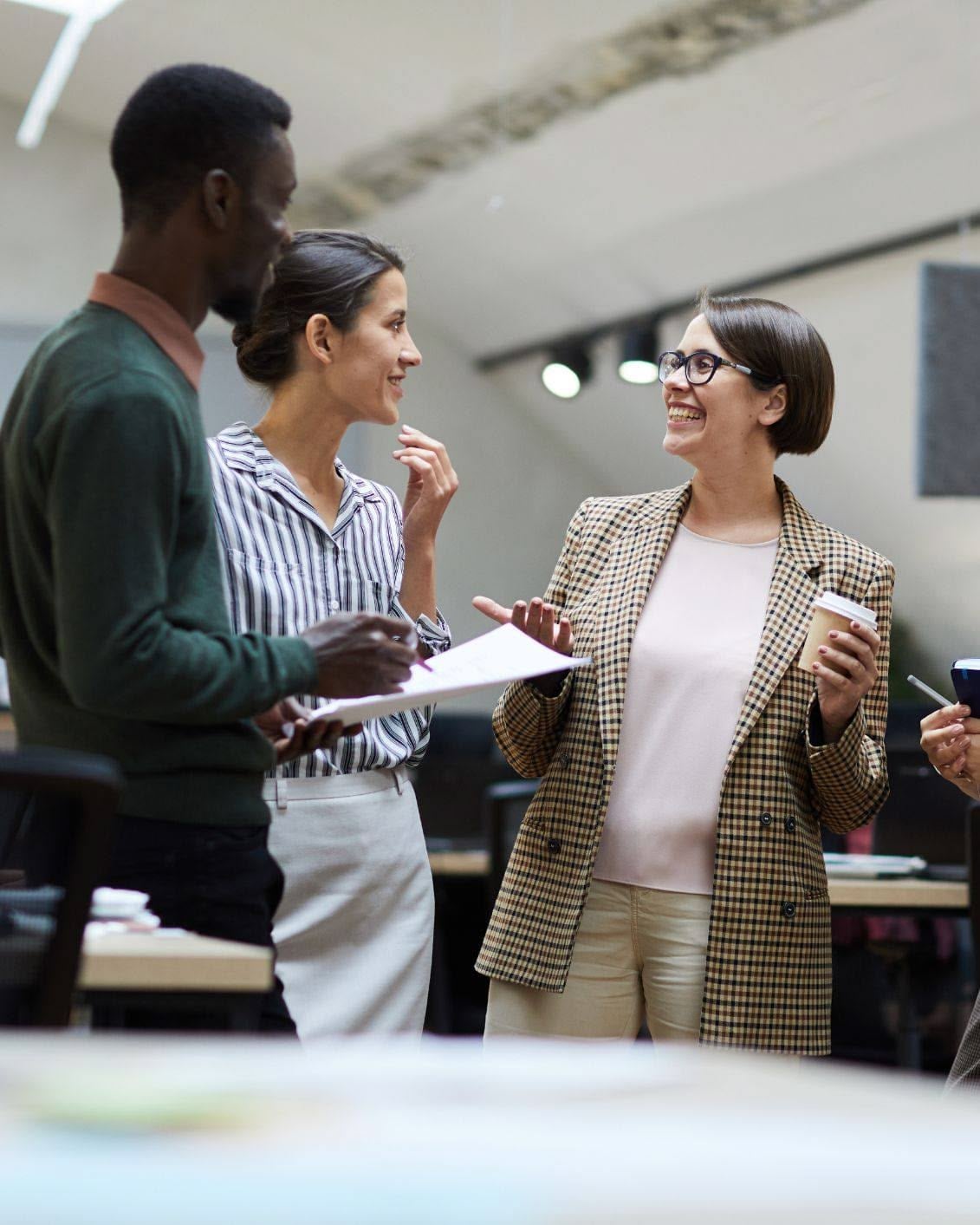 Three people stand together in an office, smiling and conversing. One holds a notepad, another a coffee cup. They appear to be engaged in a friendly discussion or meeting.