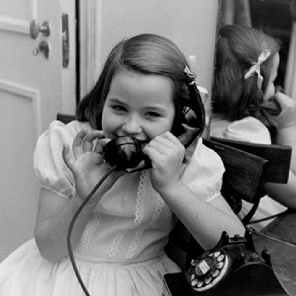 A young girl in a dress sits at a table, smiling playfully while holding a rotary phone receiver to her ear and mouth. A mirror behind her reflects her image.