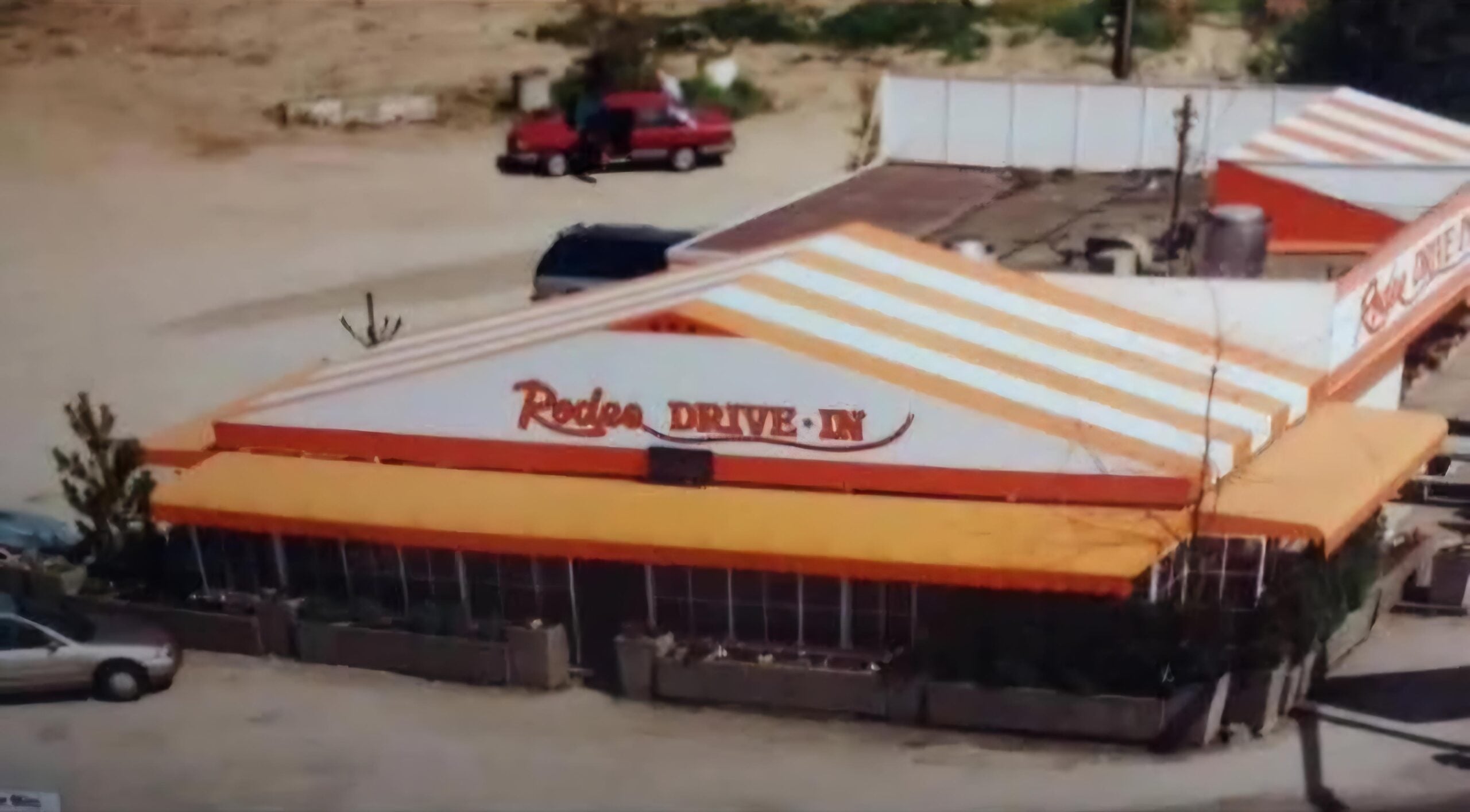 Aerial view of a retro-style diner with a white and orange striped roof, labeled "Rodeo DRIVE-IN" in red letters. Two red vehicles are parked in the background on a dirt lot.