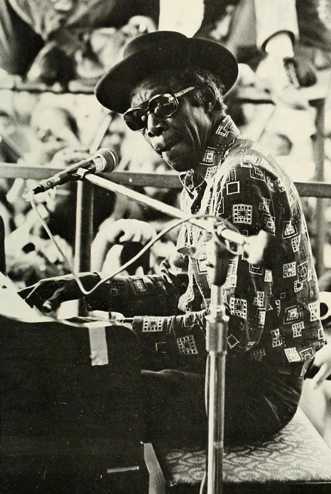 A man wearing sunglasses, a patterned shirt, and a hat plays the piano and sings into a microphone at an outdoor event, with a crowd visible in the background. The image is in black and white.
