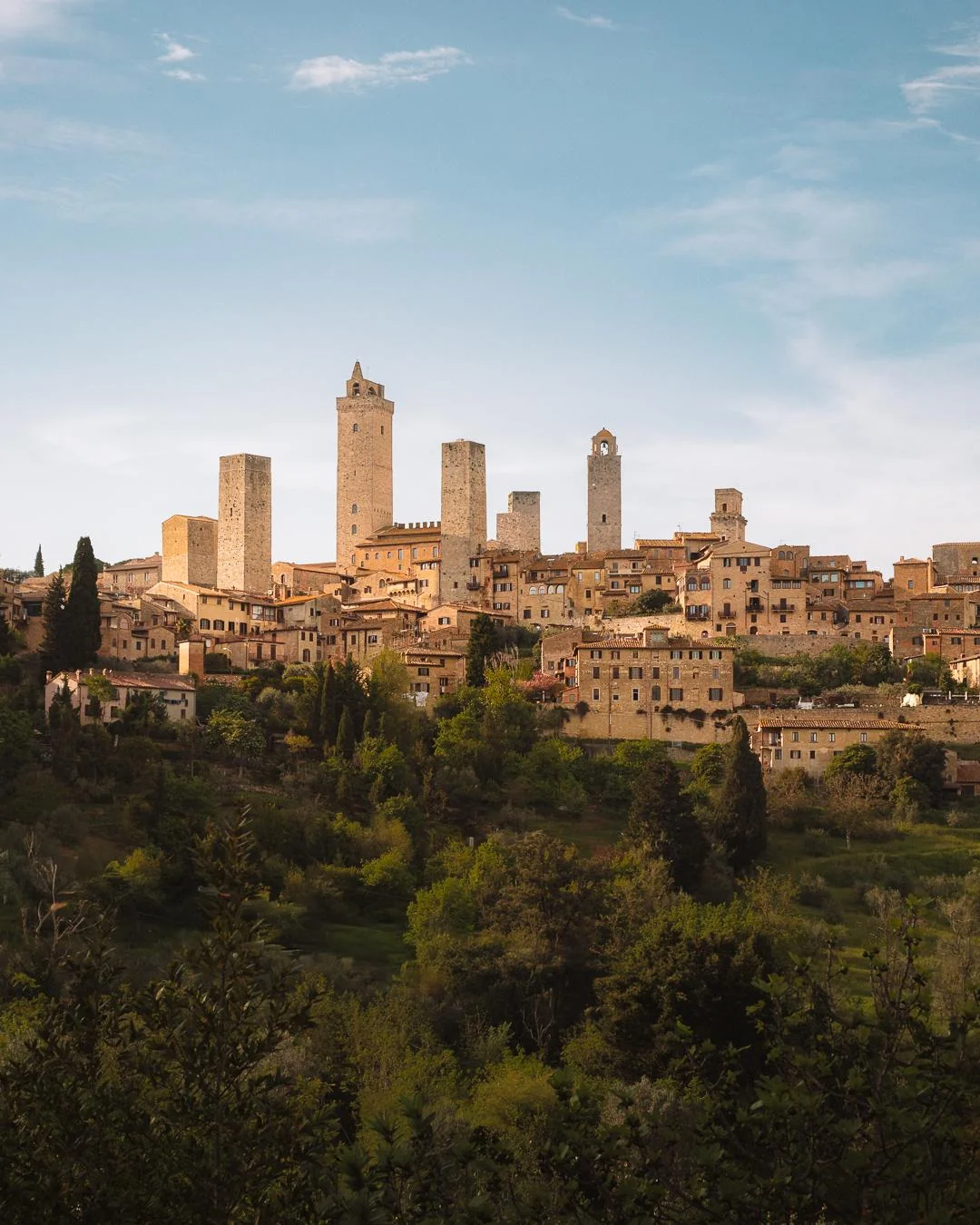 A scenic view of San Gimignano, Italy, showing medieval stone towers rising above rustic buildings with tiled roofs, set against a blue sky and surrounded by green trees and countryside.