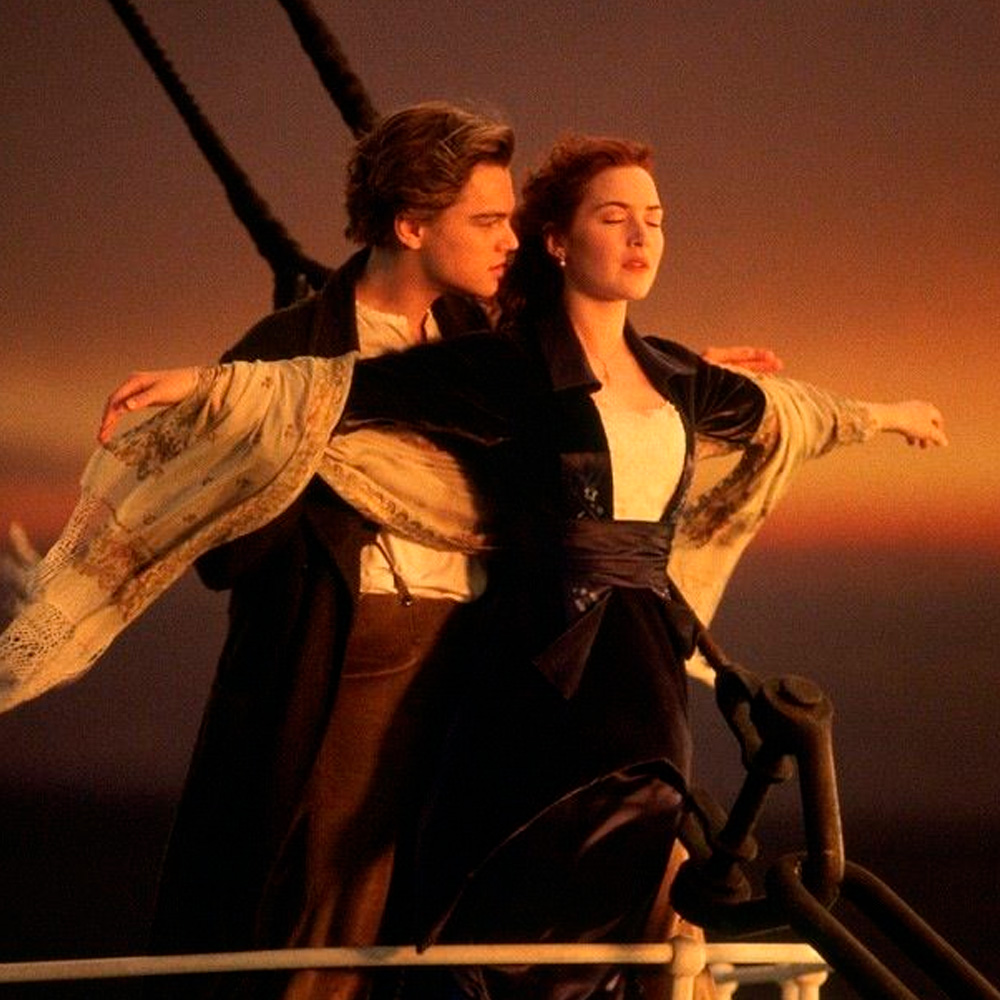 Two people stand at the bow of a ship at sunset, with one person spreading their arms wide and the other standing closely behind, gently holding them, recreating an iconic scene from the film "Titanic.