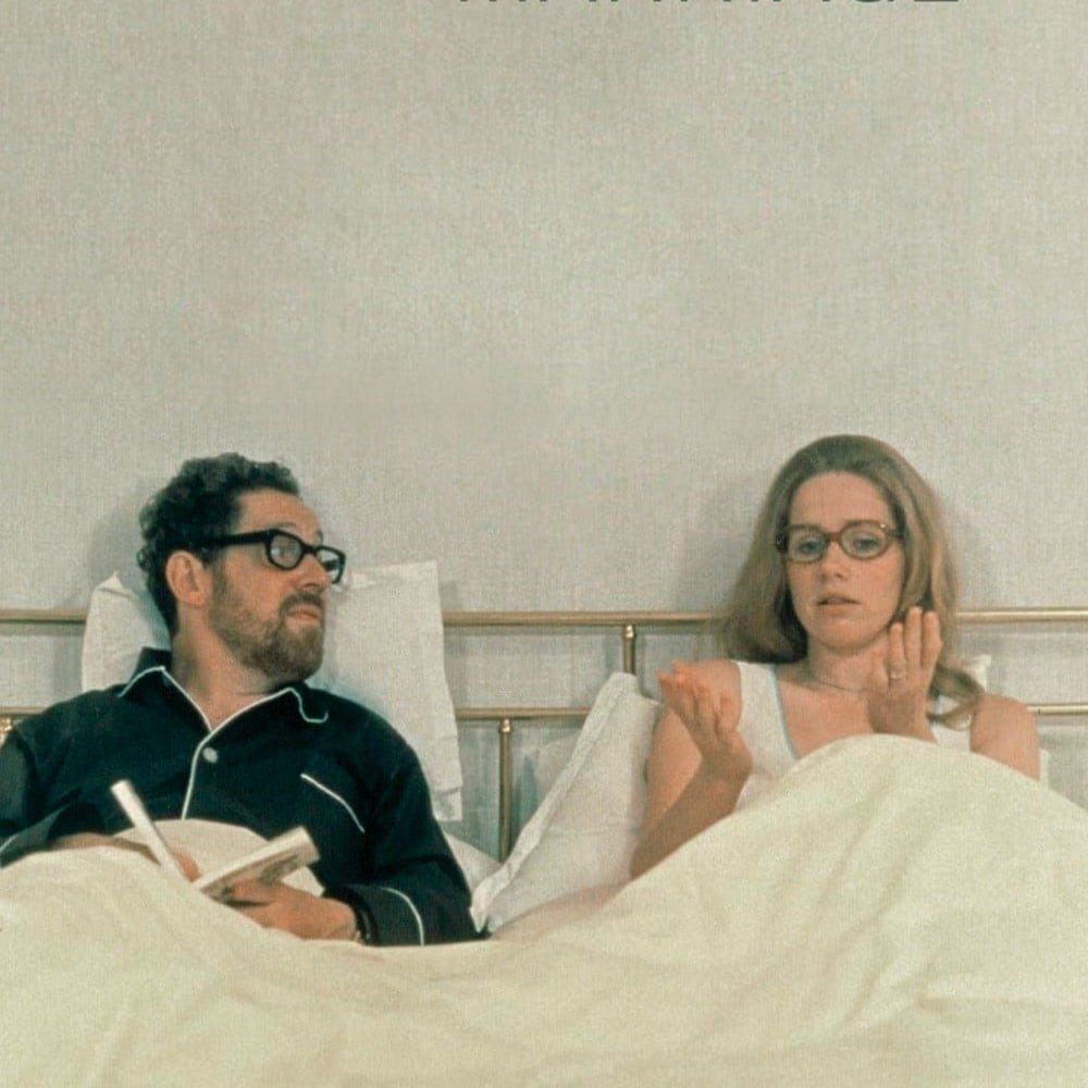 A man and woman with glasses sit up in bed under white covers. The man is holding a book and looking at the woman, while the woman gestures with her hands, appearing to explain something.