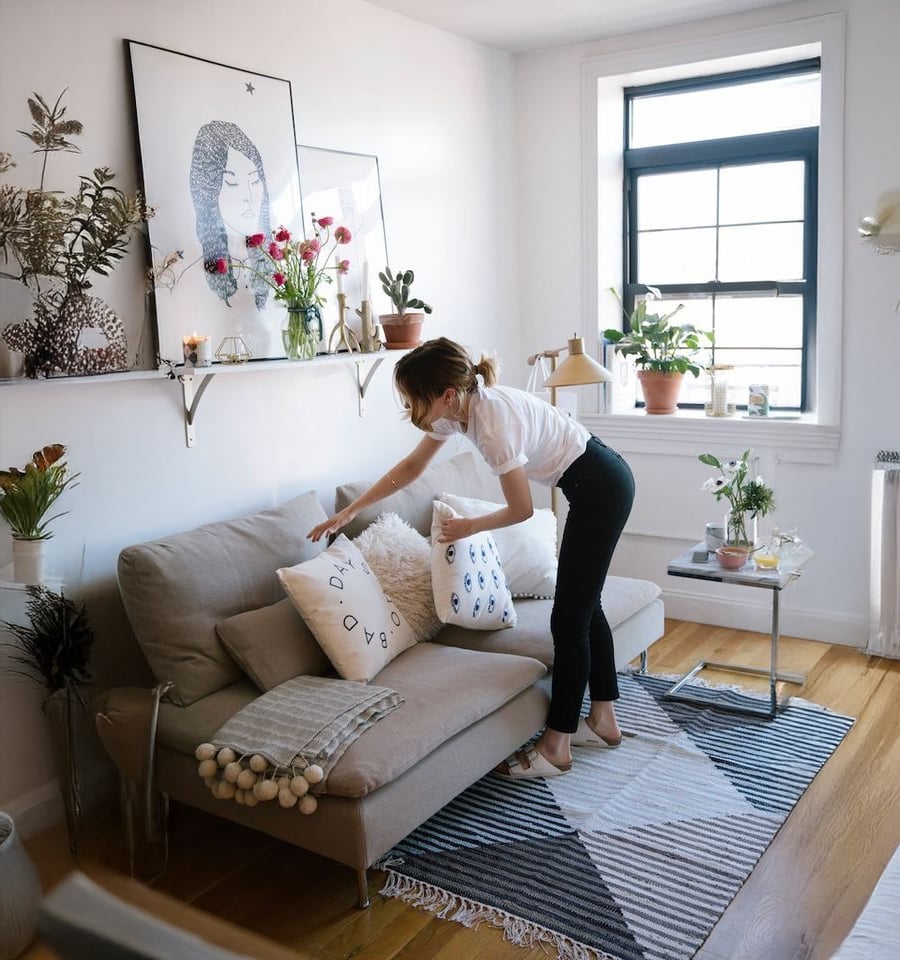 A woman arranges pillows on a beige sofa in a bright, modern living room with wooden floors, plants, a window, wall art, and decorative shelves. A patterned rug and throw add cozy touches to the space.