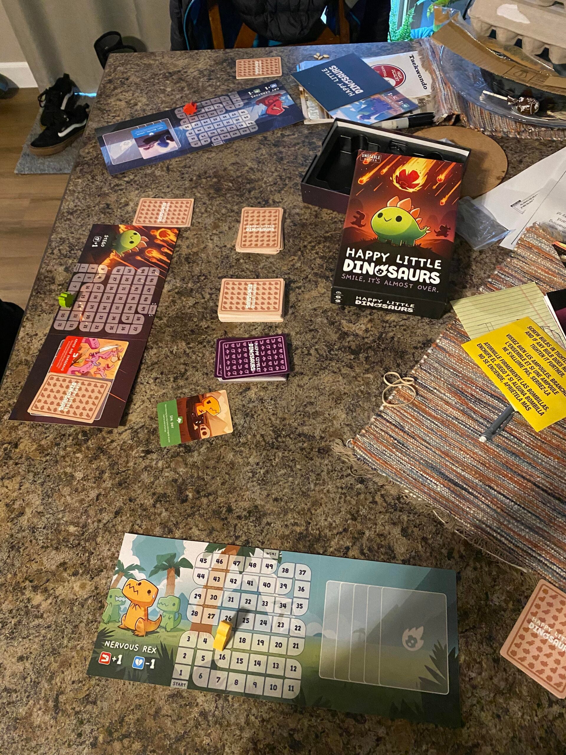 A tabletop with the board game "Happy Little Dinosaurs" set up, showing player boards, cards, and game pieces scattered on a brown counter among papers and personal items.