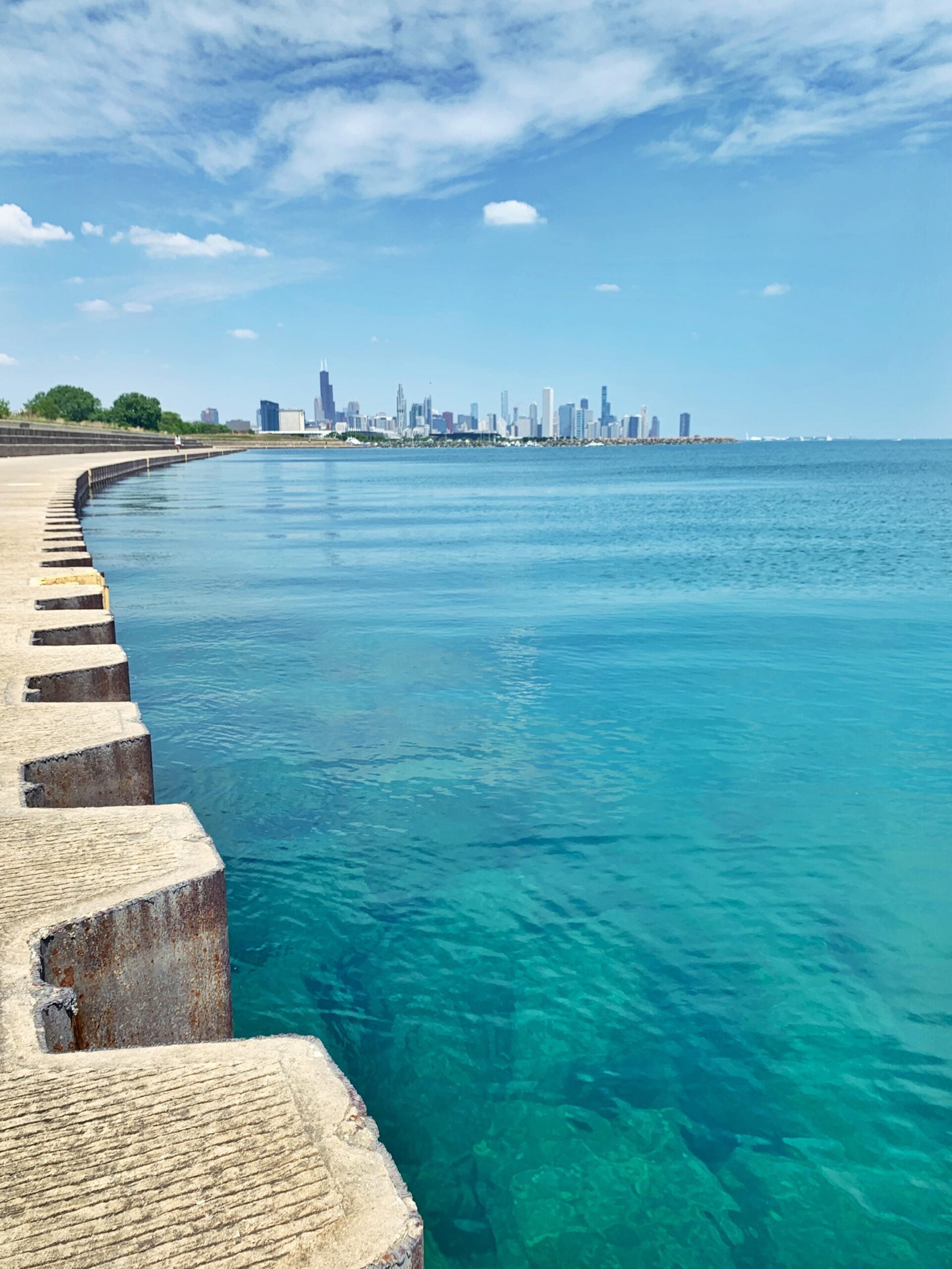 A clear blue lake meets a concrete shoreline with a distant city skyline under a partly cloudy sky. The water is calm, and the city buildings rise on the horizon.