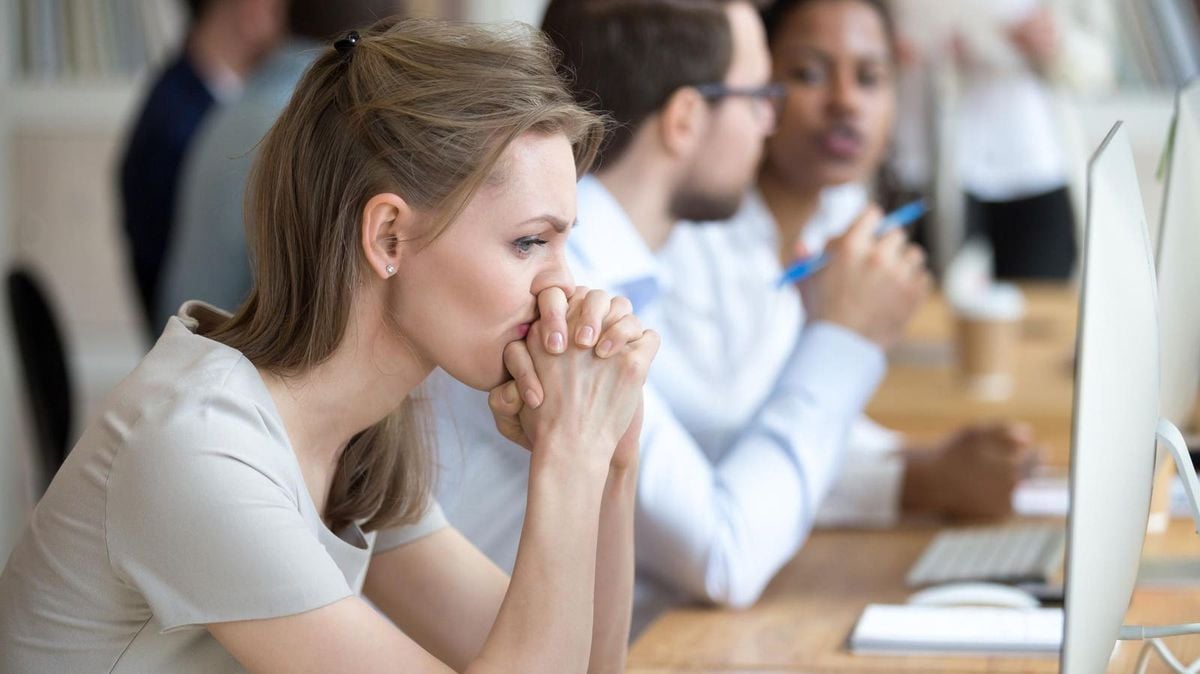 A woman sits at a desk, looking intently at a computer screen with her hands clasped near her face. Other people work at computers beside her in a modern office setting.